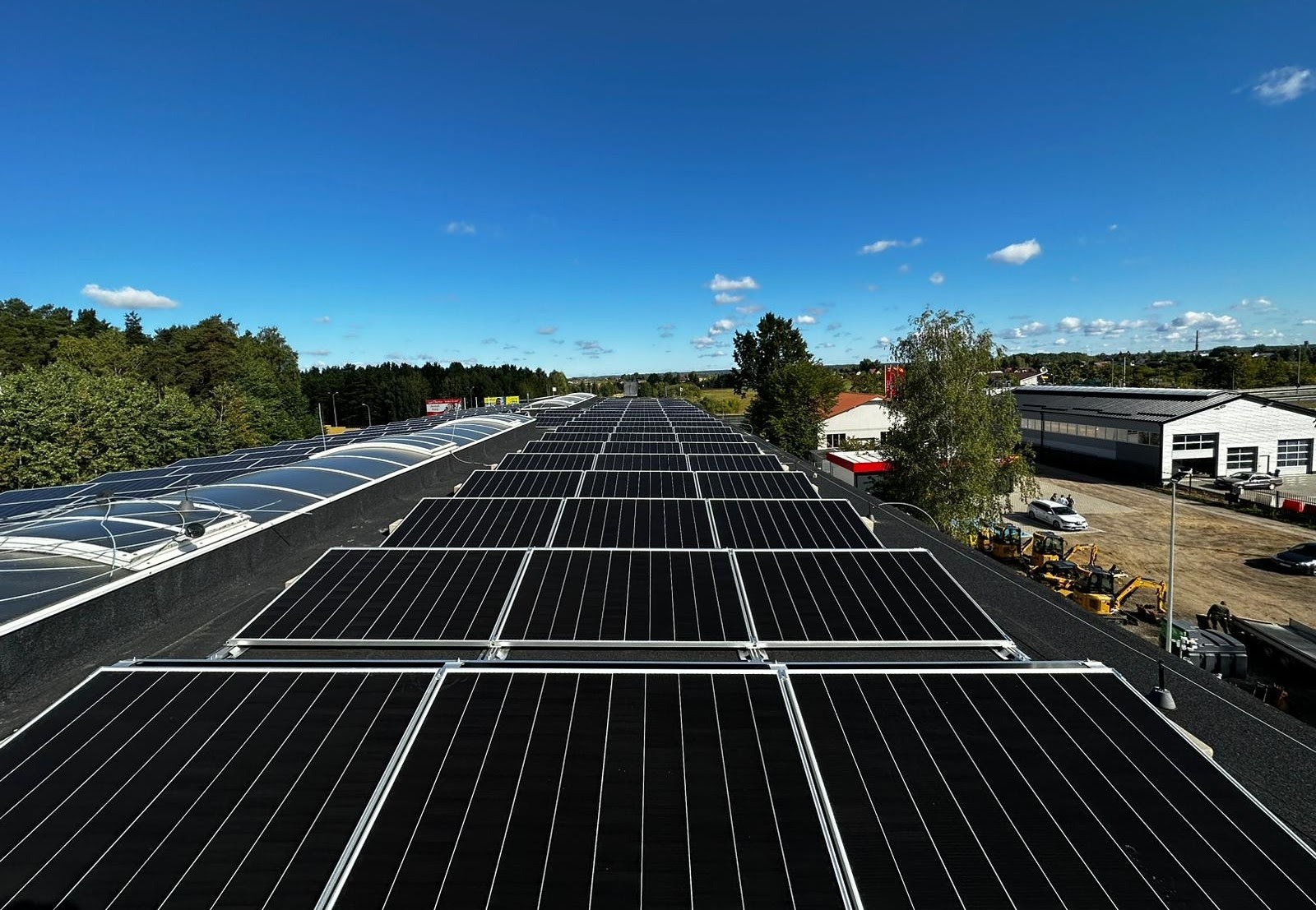 Widok z góry na dużą instalację paneli słonecznych na płaskim dachu budynku przemysłowego, z zielonymi drzewami i błękitnym niebem w tle.