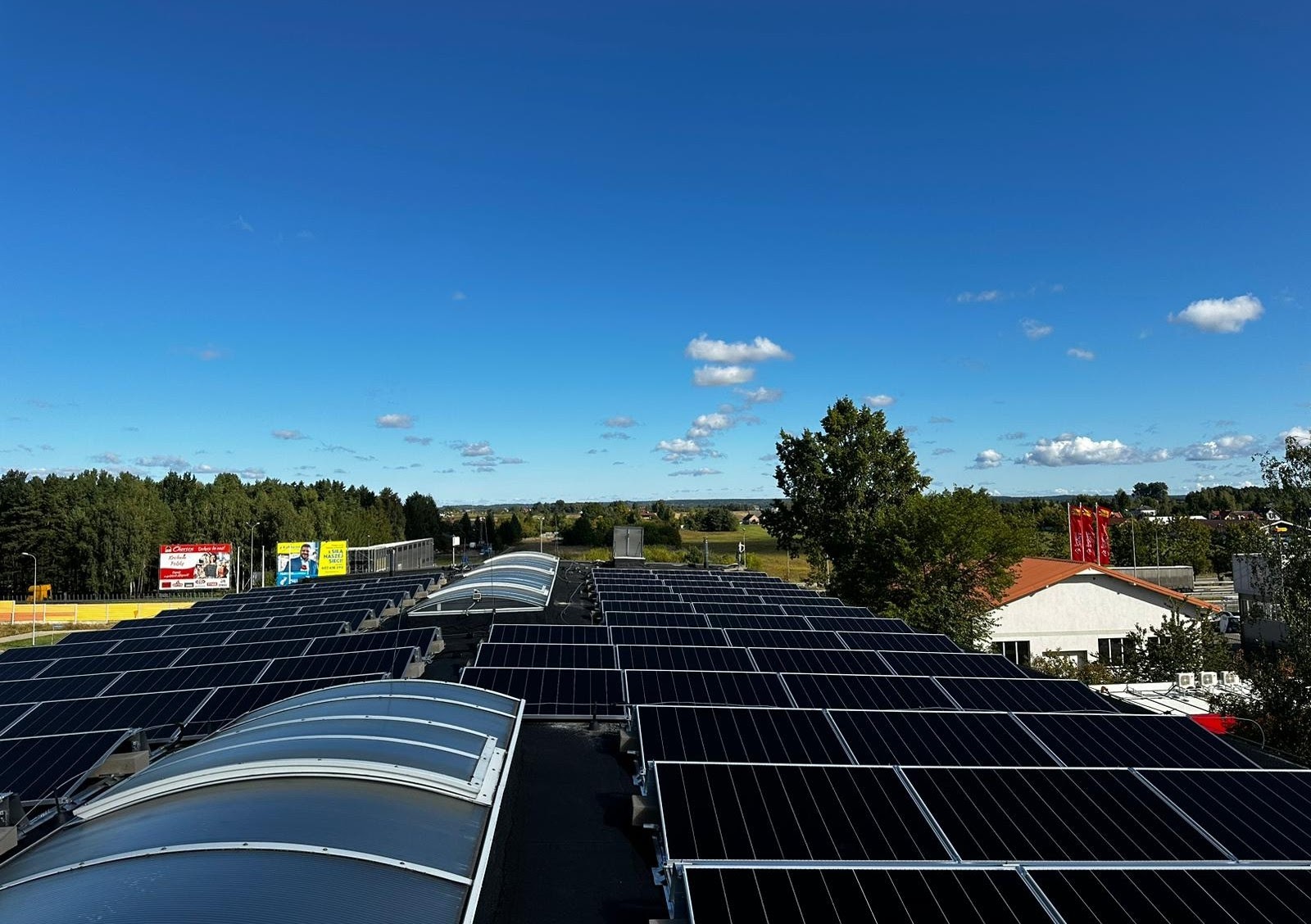 Widok z góry na dużą instalację paneli słonecznych na dachu budynku z widocznymi świetlikami, w tle drzewa i błękitne niebo z chmurami.