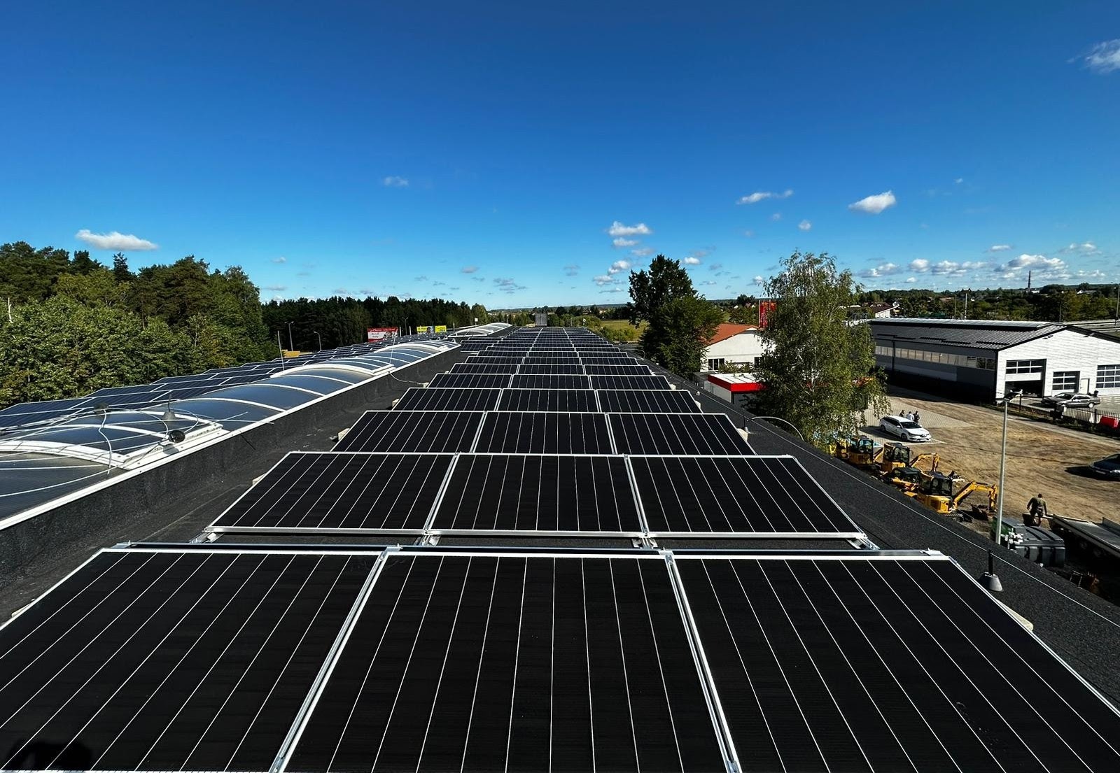 Rozległa instalacja fotowoltaiczna na dachu budynku przemysłowego w słoneczny dzień, z widocznymi maszynami budowlanymi w tle.