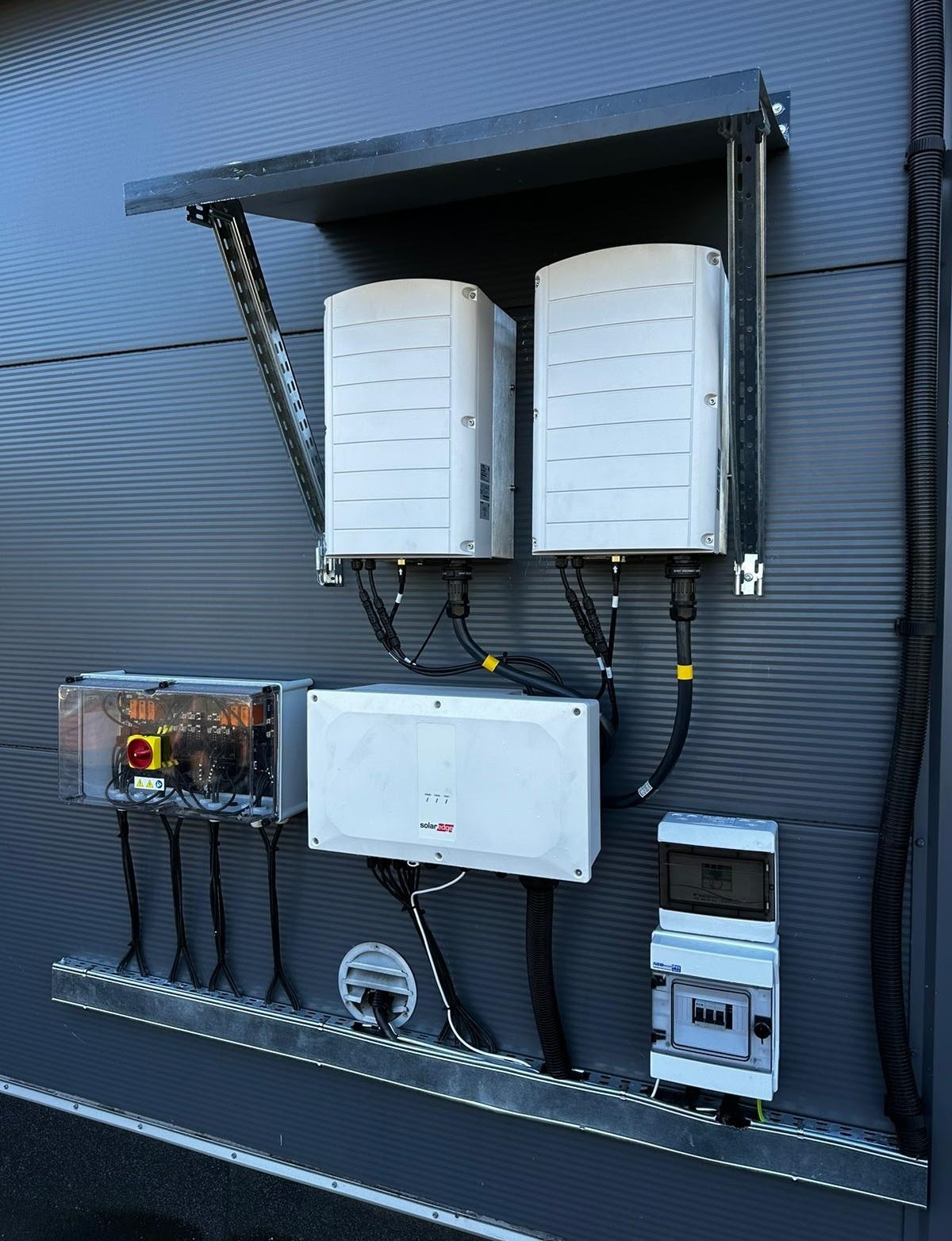 Dwa białe falowniki zamontowane na szarej ścianie budynku, pod daszkiem ochronnym, z widocznymi kablami i skrzynkami elektrycznymi.