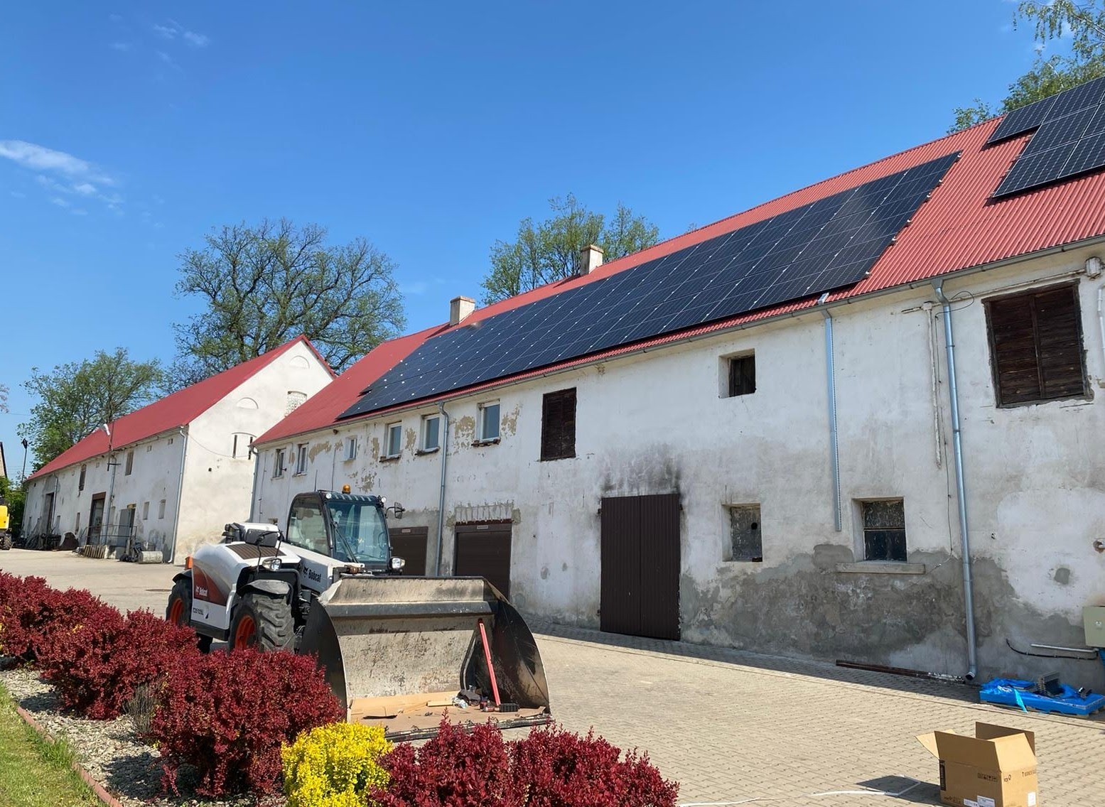 Montaż paneli słonecznych na dachu starego budynku gospodarczego z czerwoną dachówką, na pierwszym planie ładowarka teleskopowa Bobcat i rabata z czerwono-żółtymi kwiatami.