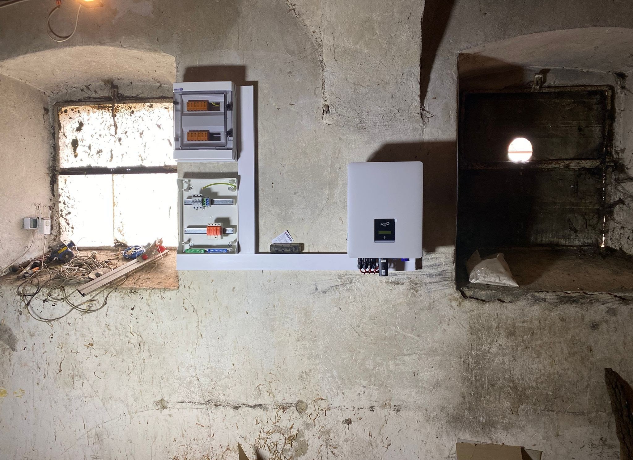 Montaż instalacji w piwnicy: inwerter FOX, skrzynka rozdzielcza i okablowanie na tle surowych, betonowych ścian z dwoma oknami.