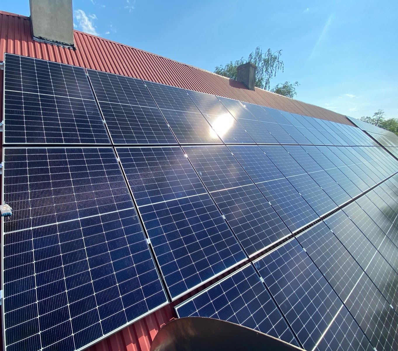 Panele słoneczne na czerwonym dachu domu jednorodzinnego, widoczne z dołu pod kątem, z refleksami słońca na powierzchni paneli.