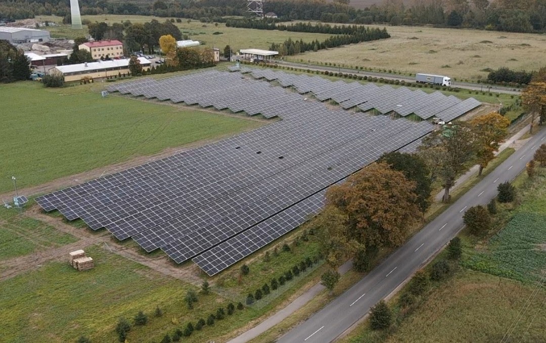 Rozległa farma paneli słonecznych w Słupsku, widok z lotu ptaka, panele ułożone rzędami na zielonym polu, obok droga i drzewa w jesiennych barwach, w tle budynki gospodarcze i las.