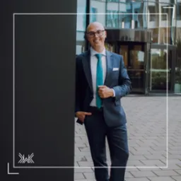 Uśmiechnięty mężczyzna w okularach i granatowym garniturze z turkusowym krawatem, stojący przed nowoczesnym biurowcem.