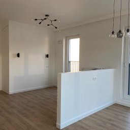 Jarbud.lux - Jasne wnętrze po remoncie: białe ściany, drewnopodobne panele, nowoczesne lampy. Widoczne gniazdka, okno z widokiem na budynek. Minimalistyczny design.