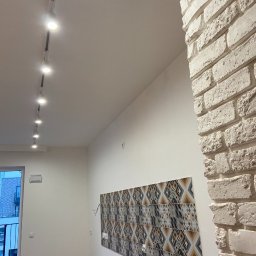 Jarbud.lux - Wykończenie wnętrza: biała cegła na rogu ściany, dekoracyjne panele ścienne z geometrycznym wzorem, oświetlenie szynowe na suficie. Nowoczesny design.