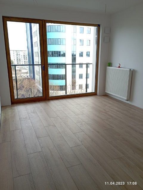 Jasne panele podłogowe w nowym mieszkaniu z dużym oknem balkonowym i widokiem na miasto, grzejnik na ścianie. Minimalistyczne wnętrze.