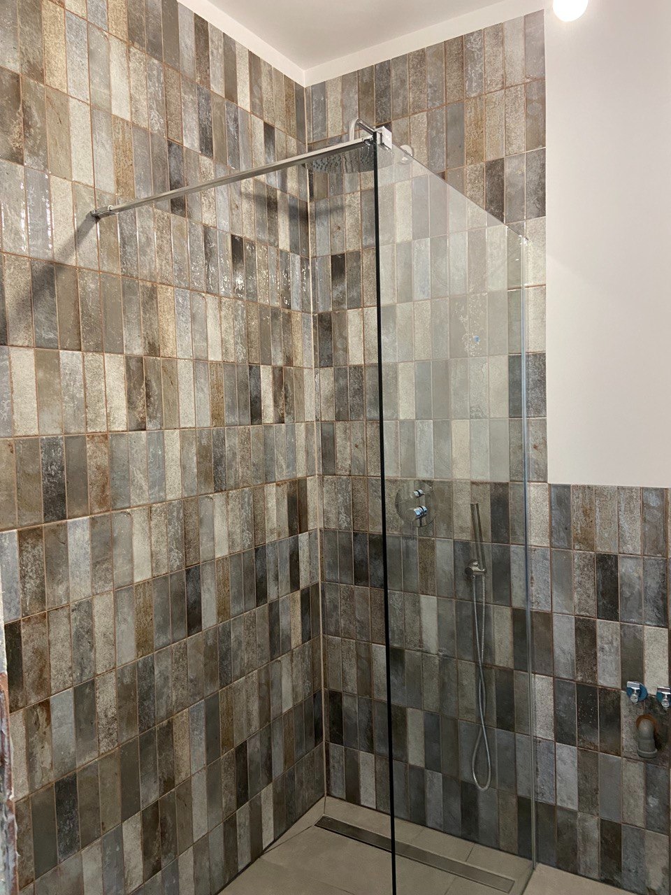Wykończona kabina prysznicowa z mozaiką w odcieniach szarości i beżu, z chromowaną armaturą i szklaną ścianką. Widoczny odpływ liniowy w podłodze.