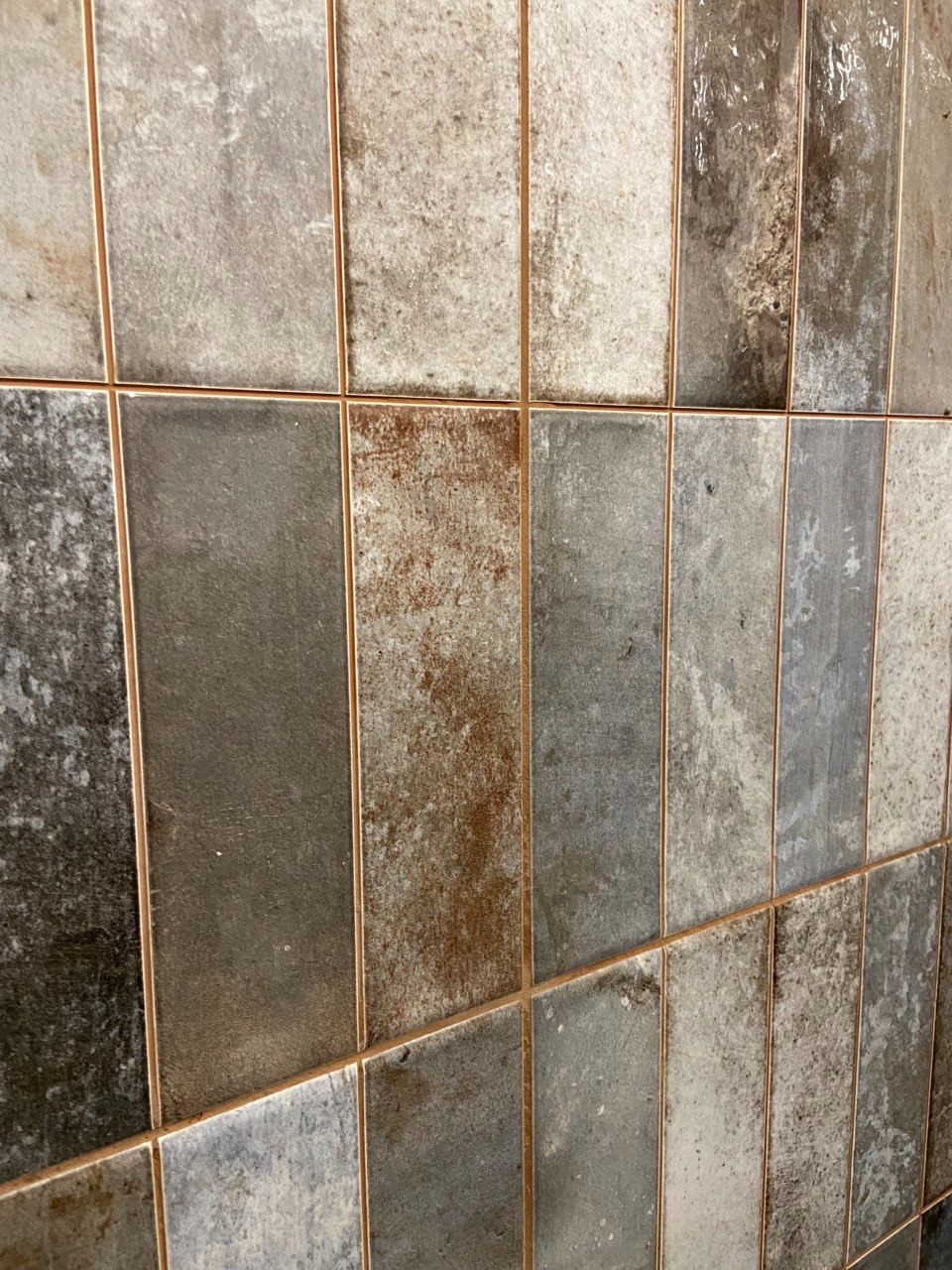 Ściana wyłożona szarymi, prostokątnymi płytkami imitującymi kamień, z wyraźnymi, złotymi fugami. Ułożenie pionowe, zróżnicowana kolorystyka płytek.