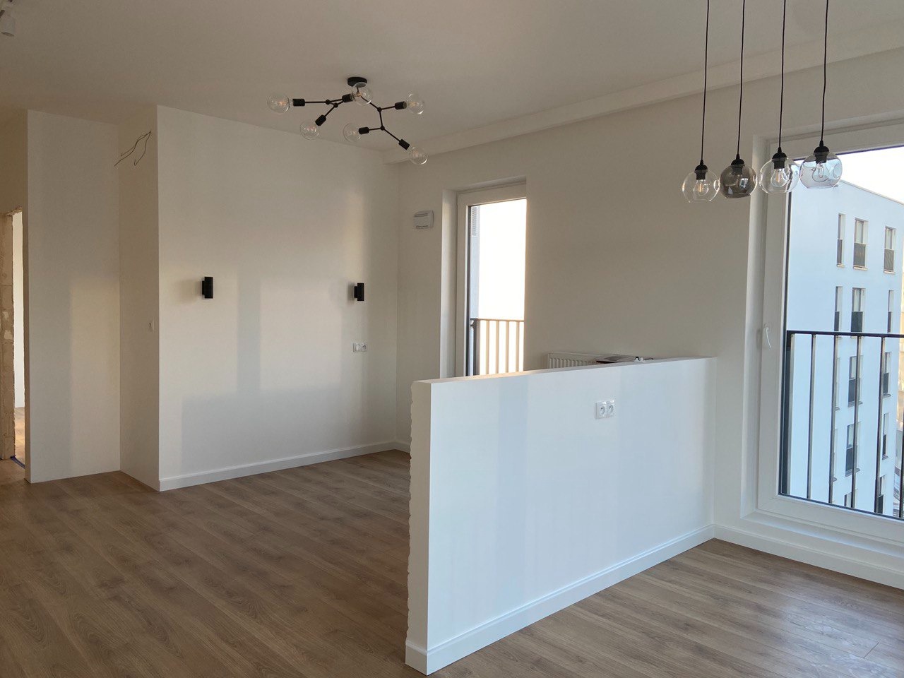 Jasne wnętrze po remoncie: białe ściany, drewnopodobne panele, nowoczesne lampy. Widoczne gniazdka, okno z widokiem na budynek. Minimalistyczny design.