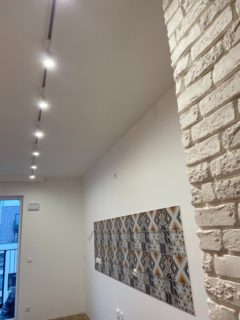 Wykończenie wnętrza: biała cegła na rogu ściany, dekoracyjne panele ścienne z geometrycznym wzorem, oświetlenie szynowe na suficie. Nowoczesny design.