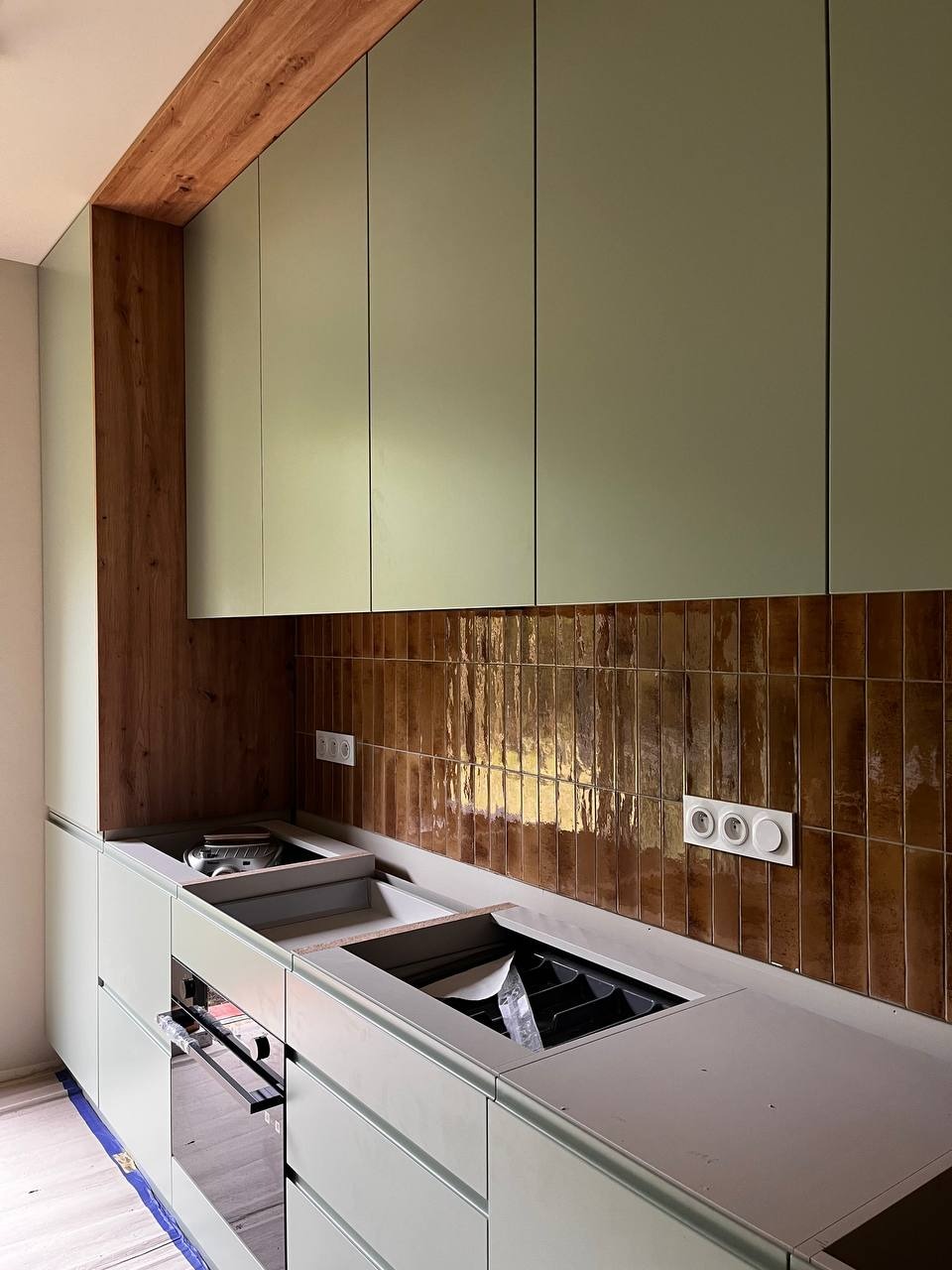 Nowoczesna kuchnia w trakcie montażu: oliwkowe szafki, brązowe płytki na ścianie, drewniane akcenty, widoczne gniazdka elektryczne. Ujęcie z bliska na elementy zabudowy.