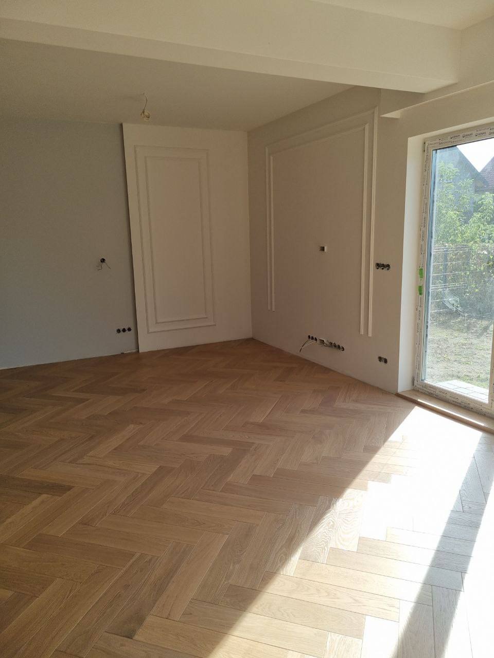 Jasne, przestronne wnętrze po remoncie: parkiet ułożony w jodełkę, białe ściany z dekoracyjnymi listwami, duże okno wpuszczające światło dzienne. Widoczne gniazdka elektryczne.