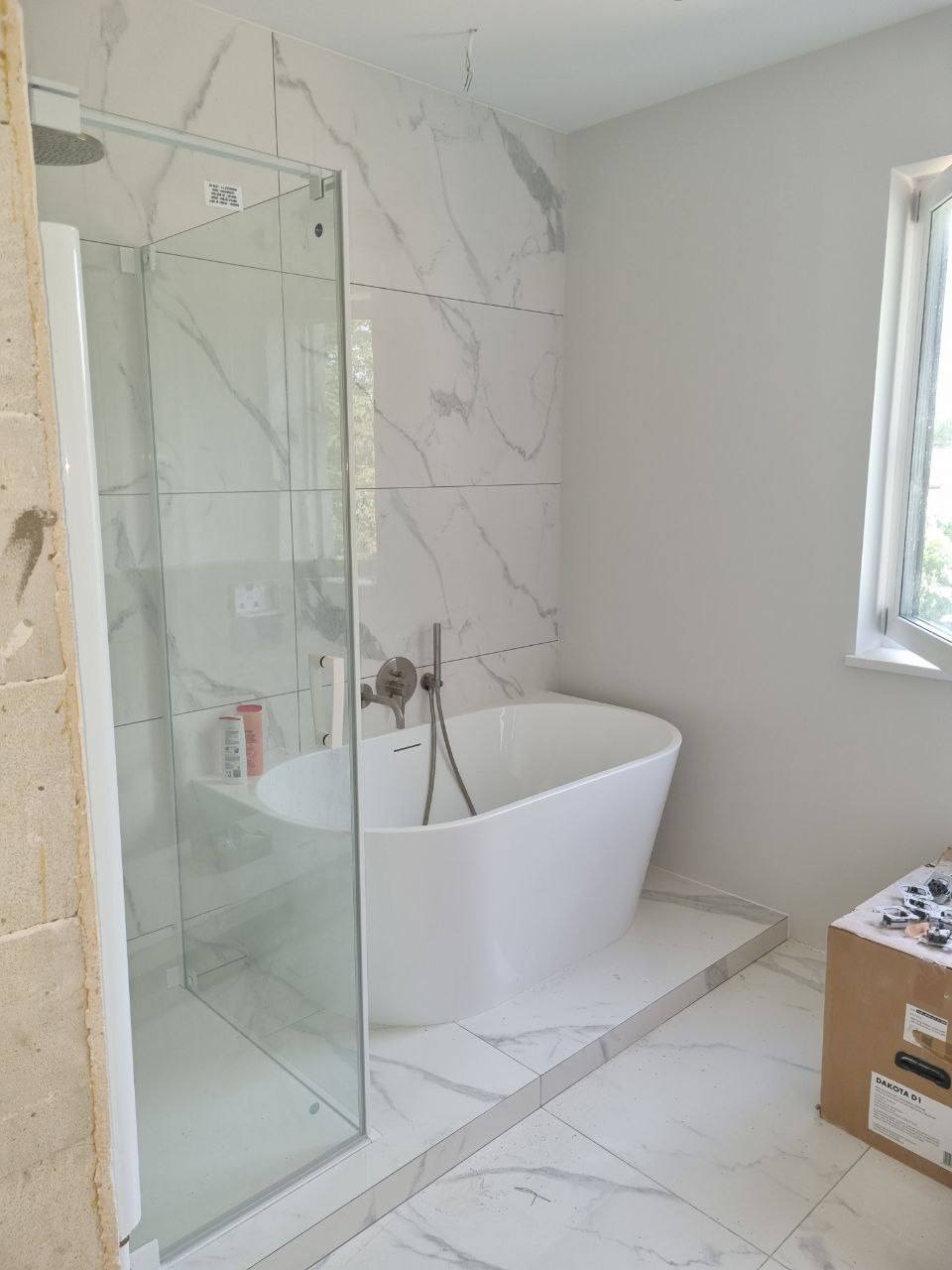 Nowoczesna łazienka z wolnostojącą wanną i kabiną prysznicową typu walk-in, wykończona płytkami imitującymi marmur, z oknem i elementami instalacji na ścianie.