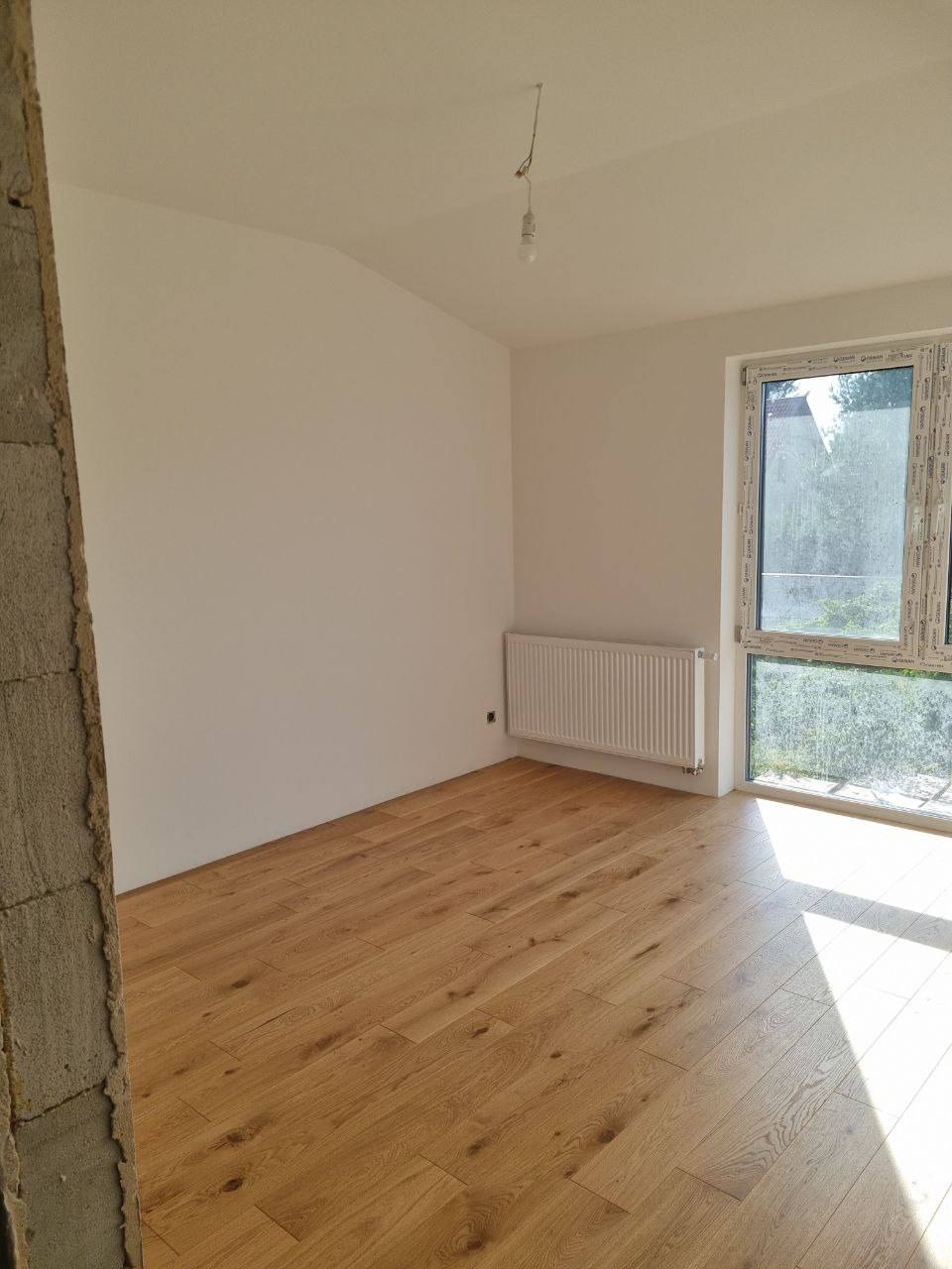 Jasne panele podłogowe w nowym pokoju z grzejnikiem pod oknem i surową ścianą. Światło słoneczne podkreśla strukturę drewna.