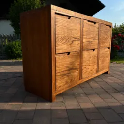 Drewniana szafka z sześcioma szufladami, wykonana z jasnego drewna z wyraźnym usłojeniem, ustawiona na brukowanym podjeździe przed domem z drewnianym płotem w tle, oświetlona popołudniowym słońcem.
