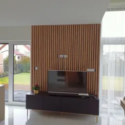 Nowoczesny salon z drewnianą ścianą z lameli, na której wisi telewizor. Pod telewizorem czarna szafka RTV na złotych nóżkach. Na szafce piloty i soundbar, obok doniczka z kwiatami. Podłoga wyłożona...