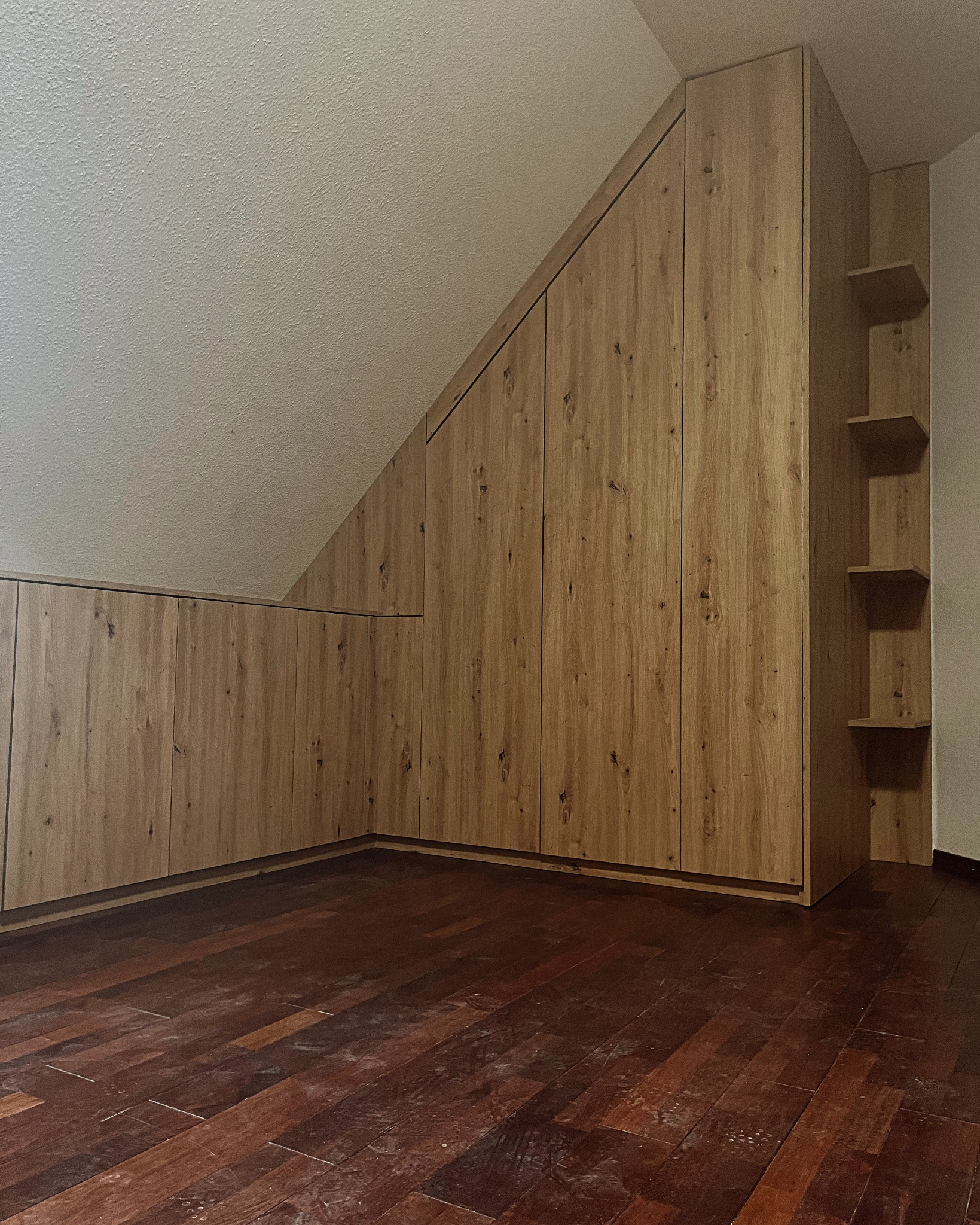 Drewniana zabudowa szafy wnękowej na poddaszu ze skośnym sufitem, z widocznymi półkami po prawej stronie, na tle ciemnej, lakierowanej podłogi parkietowej.