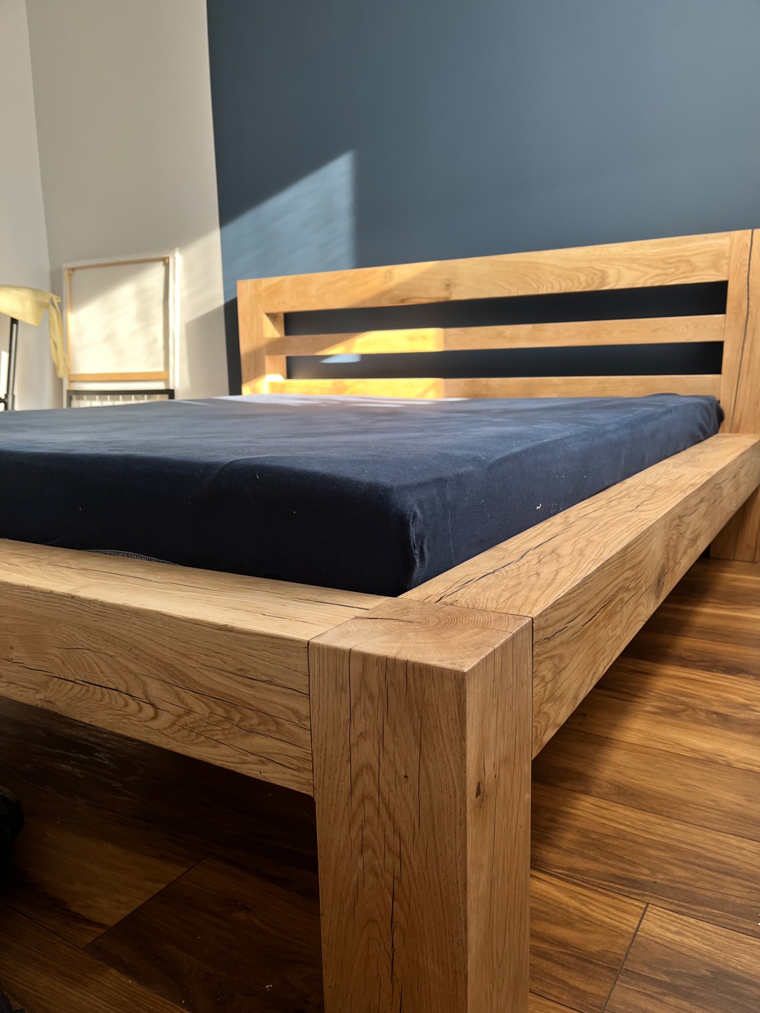 Drewniane łóżko z ciemnoniebieskim materacem, widoczne usłojenie drewna i fragment podłogi z paneli