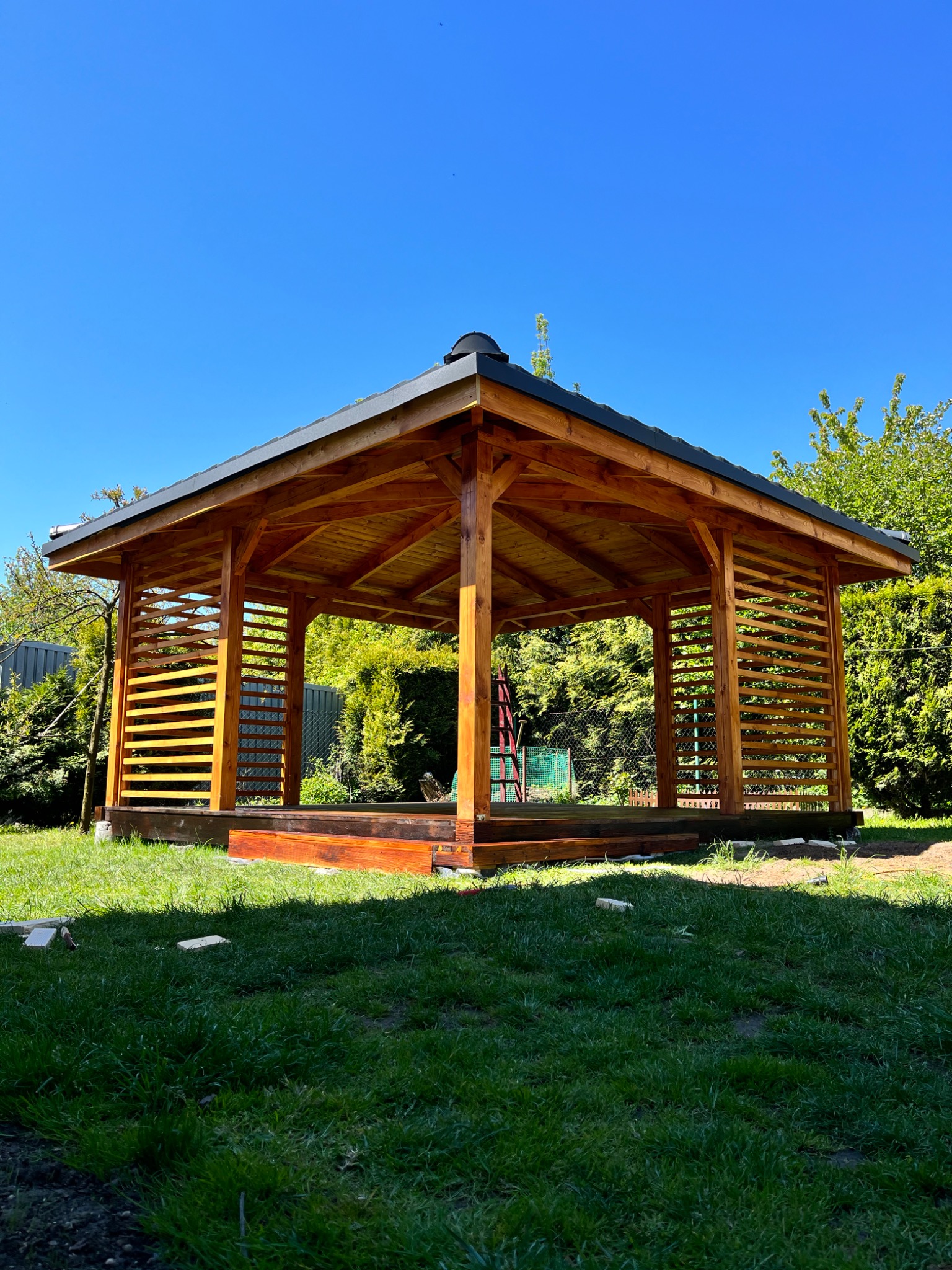 Drewniana konstrukcja altany ogrodowej z ciemnym dachem i ażurowymi ścianami, widok z dołu na tle zieleni i błękitnego nieba.