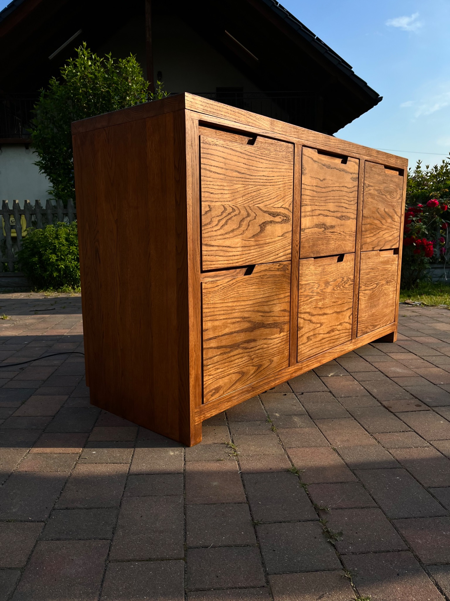 Drewniana szafka z sześcioma szufladami, wykonana z jasnego drewna z wyraźnym usłojeniem, ustawiona na brukowanym podjeździe przed domem z drewnianym płotem w tle, oświetlona popołudniowym słońcem.