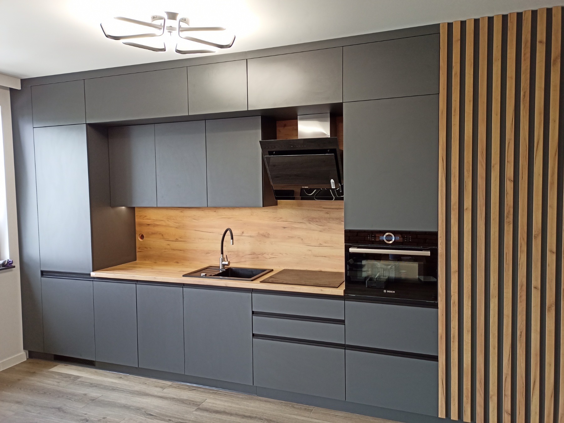 Nowoczesna kuchnia w odcieniach szarości z drewnianymi akcentami: zabudowa meblowa od podłogi do sufitu, drewniany panel dekoracyjny z pionowych listew, blat z drewnopodobnym dekorem, czarny zlew...
