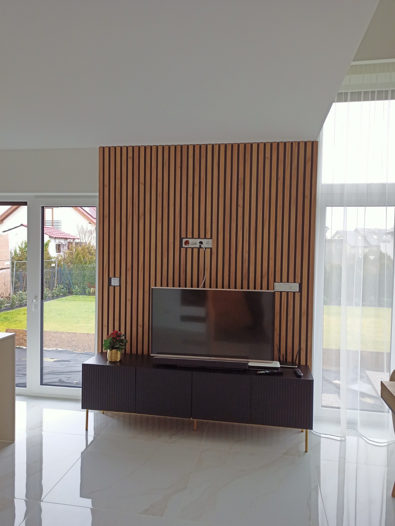 Nowoczesny salon z drewnianą ścianą z lameli, na której wisi telewizor. Pod telewizorem czarna szafka RTV na złotych nóżkach. Na szafce piloty i soundbar, obok doniczka z kwiatami. Podłoga wyłożona...