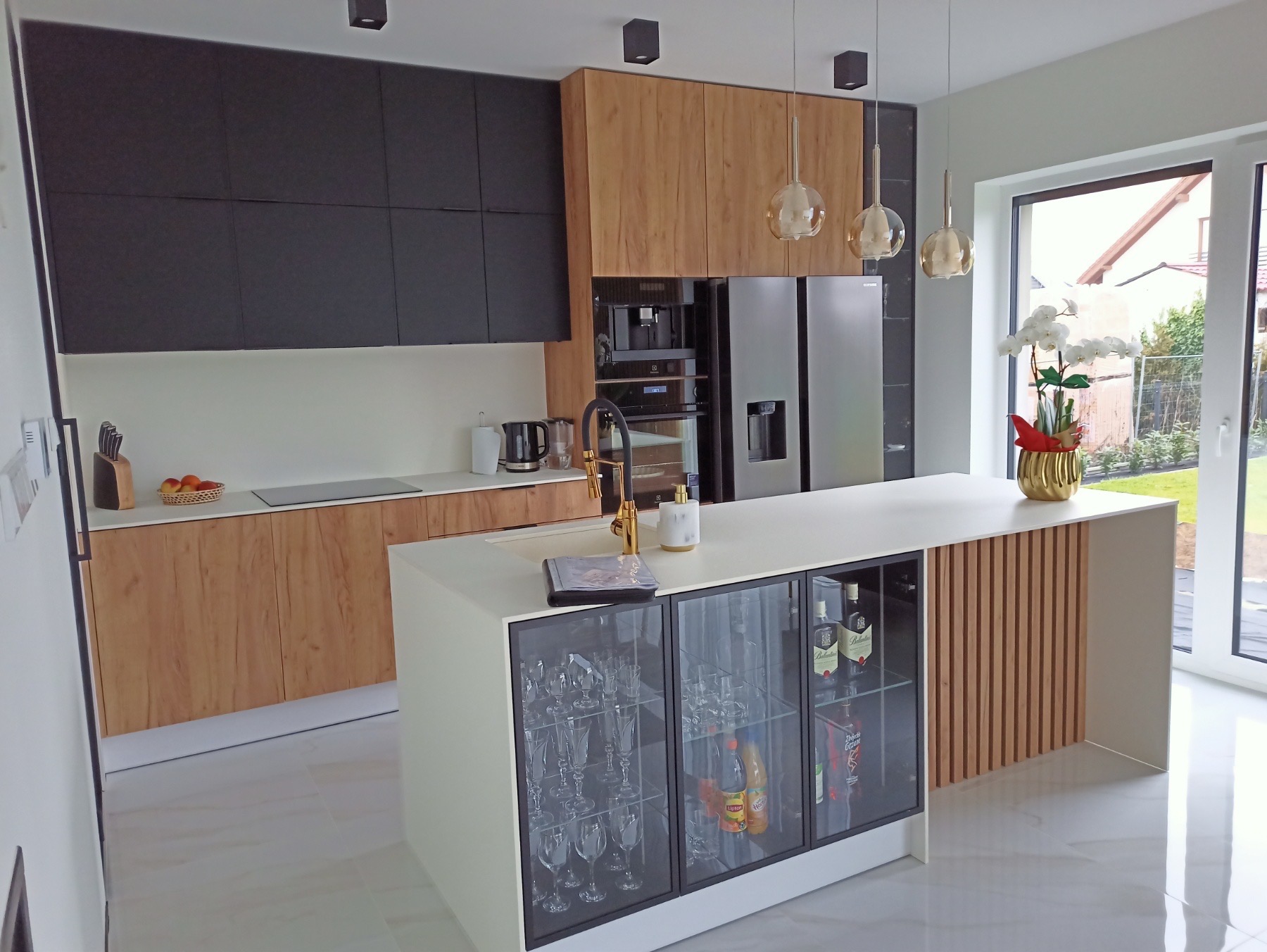 Nowoczesna kuchnia z czarnymi i drewnianymi szafkami, wyspą z przeszklonym barkiem na alkohole i złotą armaturą, oświetlona wiszącymi lampami.