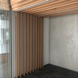 Roman Berehovyi - Nowoczesne wykończenie wnętrza biura z drewnianymi lamelami na suficie i ścianie, szara ściana z efektem betonu, ciemna podłoga i przeszklenie.