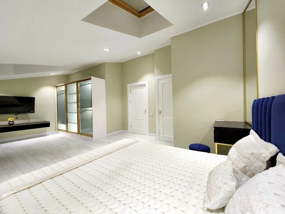 Jasna sypialnia z kremowymi ścianami, białymi drzwiami i podłogą, niebieskim zagłówkiem łóżka i oknem dachowym w suficie.