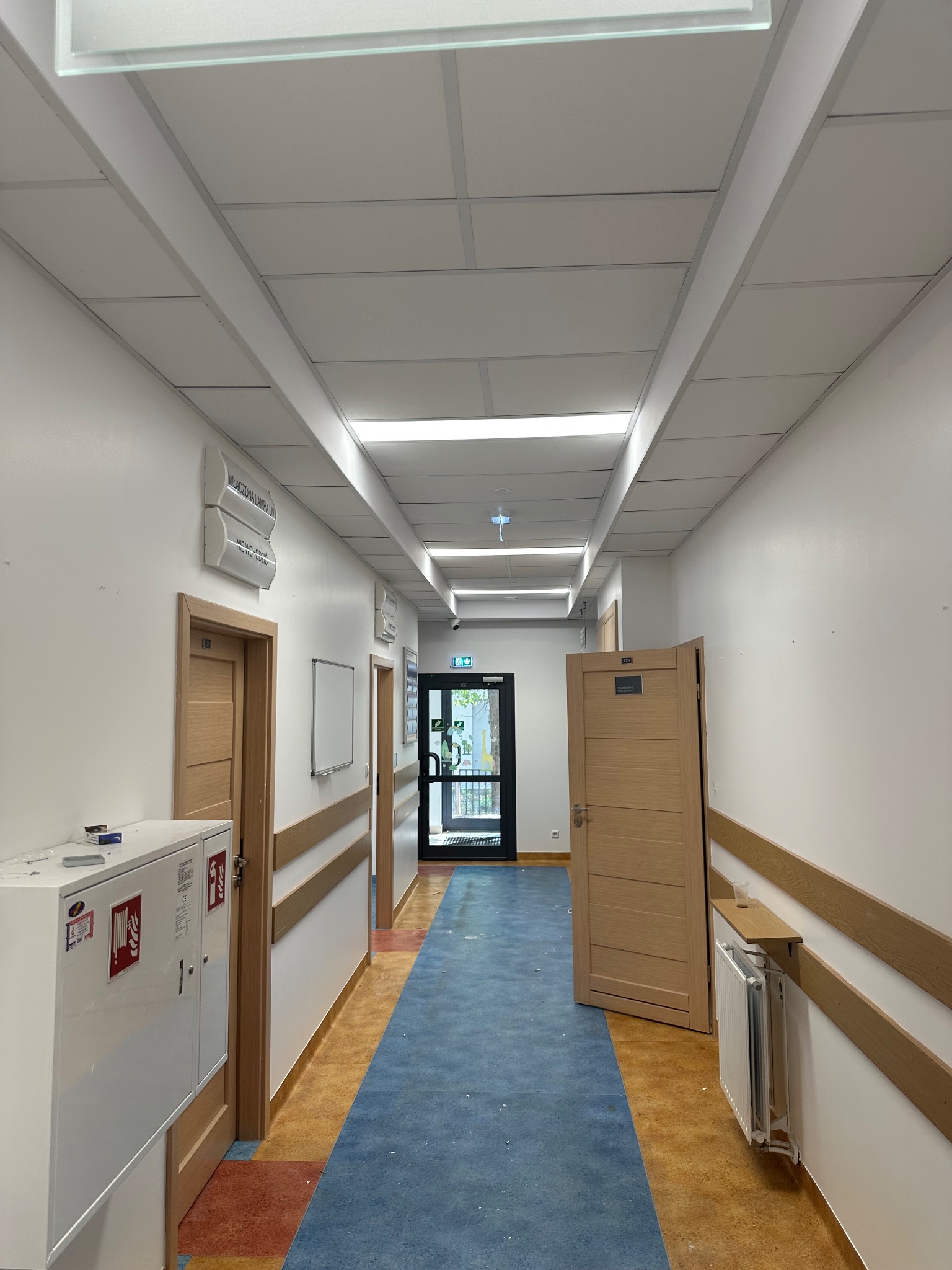Korytarz biurowy po odświeżeniu: jasne ściany, sufit podwieszany z oświetleniem, drewniane drzwi z numerami, niebieska wykładzina podłogowa.