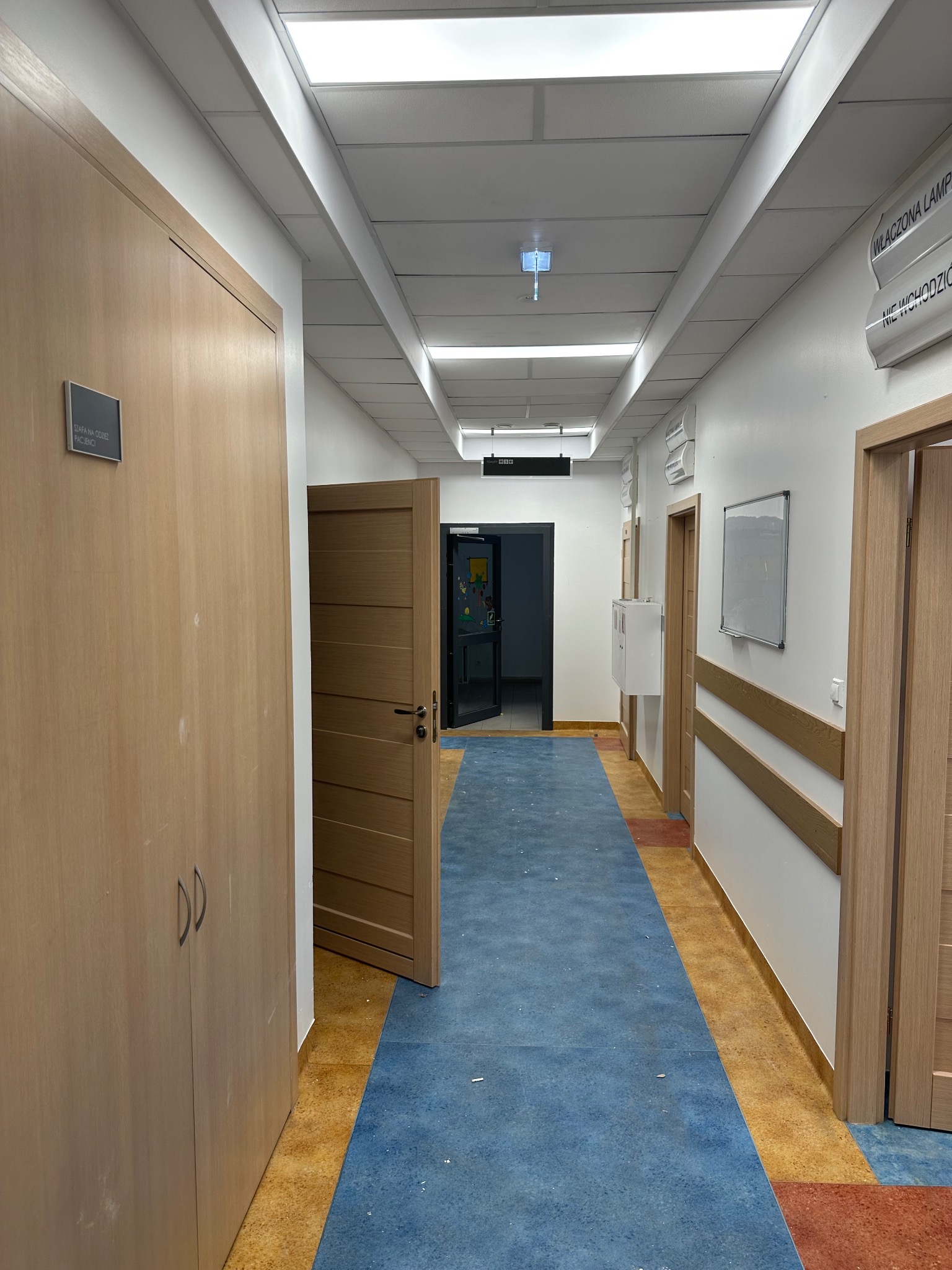 Korytarz w placówce medycznej z białymi ścianami, drewnianymi drzwiami i niebiesko-pomarańczową podłogą. Widoczne tabliczki informacyjne i szafka.