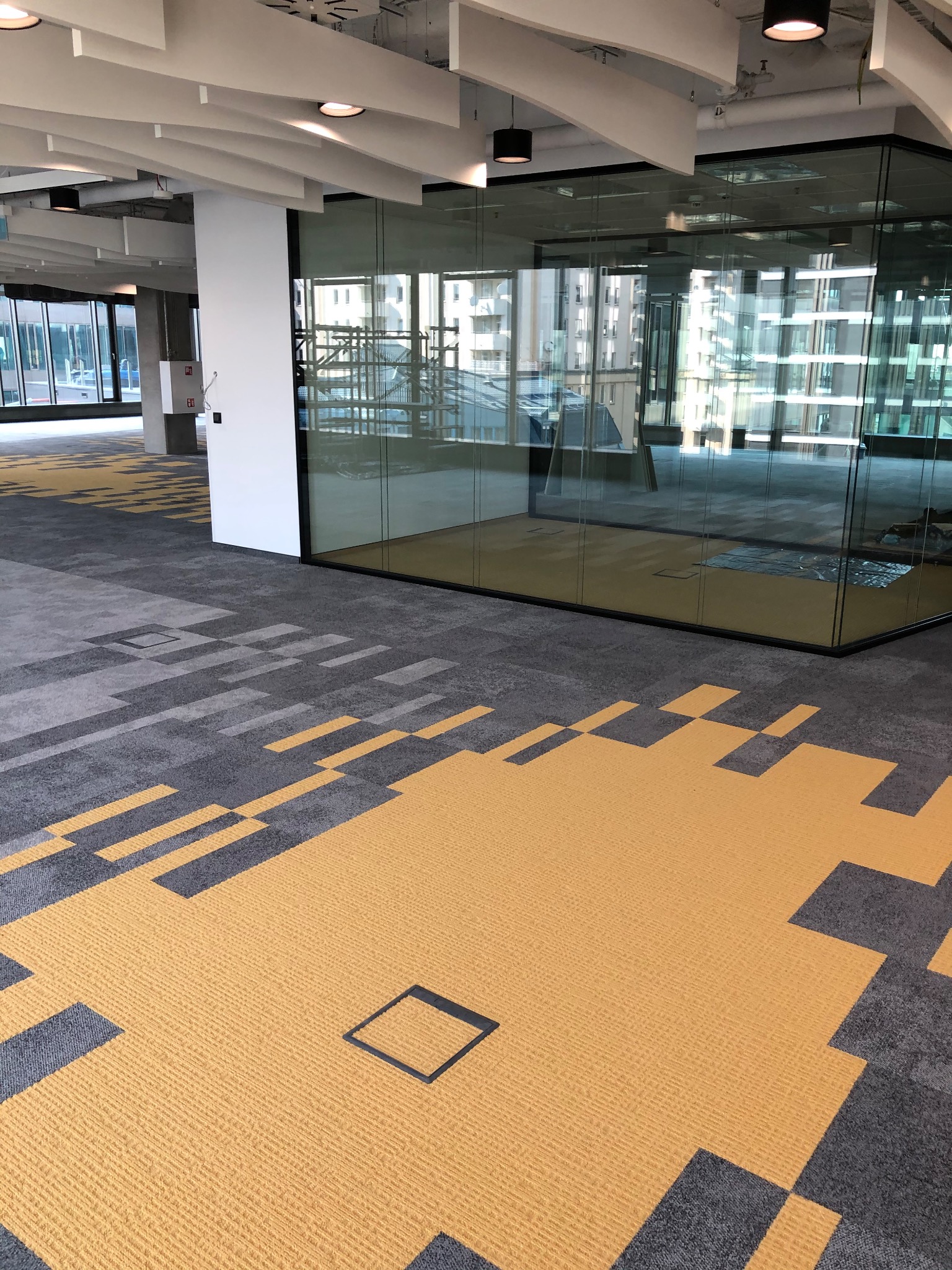 Nowoczesne biuro z żółto-szarą wykładziną dywanową w geometryczne wzory i przeszklonymi ścianami działowymi.