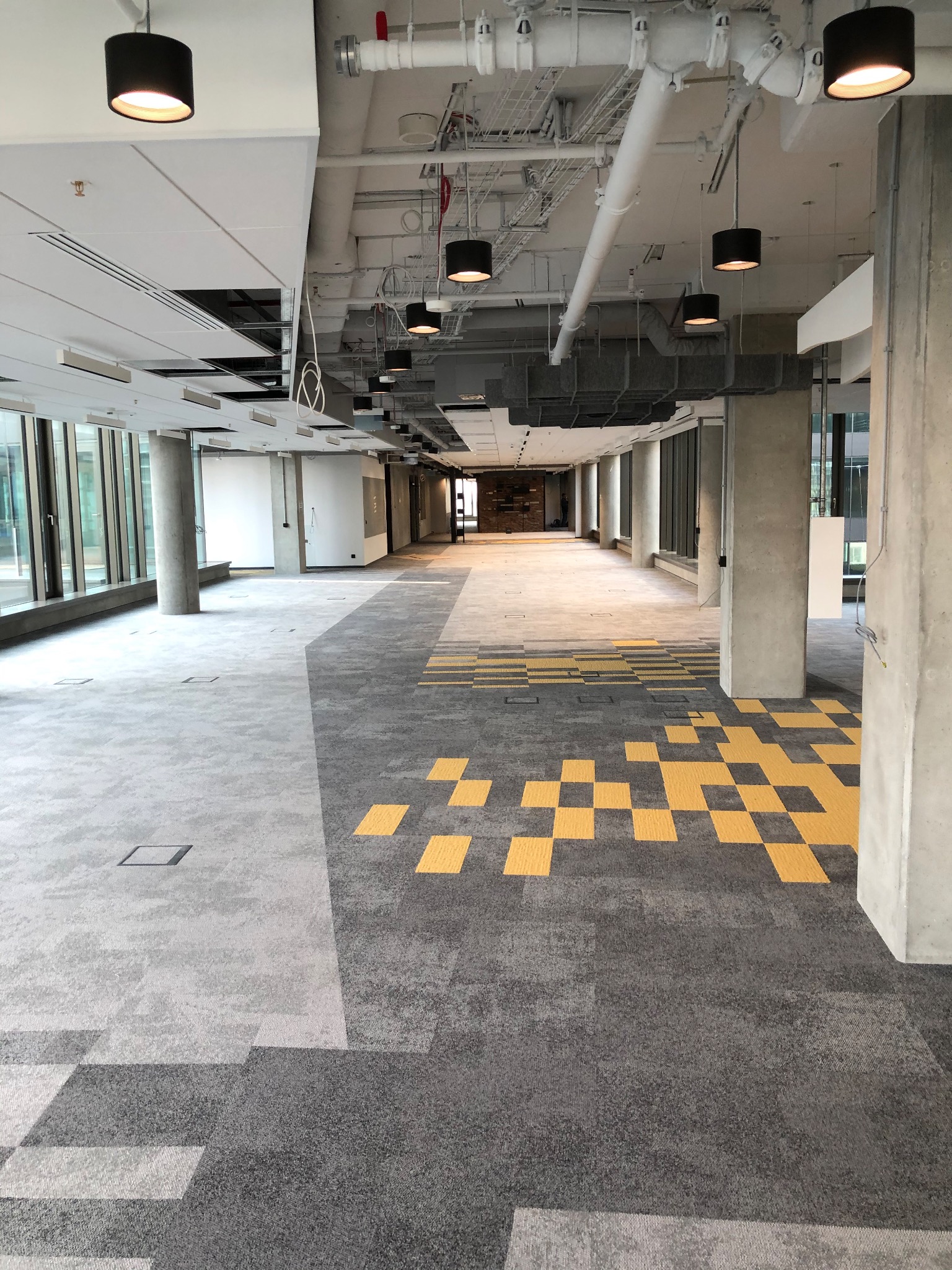 Przestronne wnętrze biurowe w stanie deweloperskim z szarą wykładziną w geometryczne wzory i żółtymi akcentami, widoczne betonowe kolumny i instalacje pod sufitem.