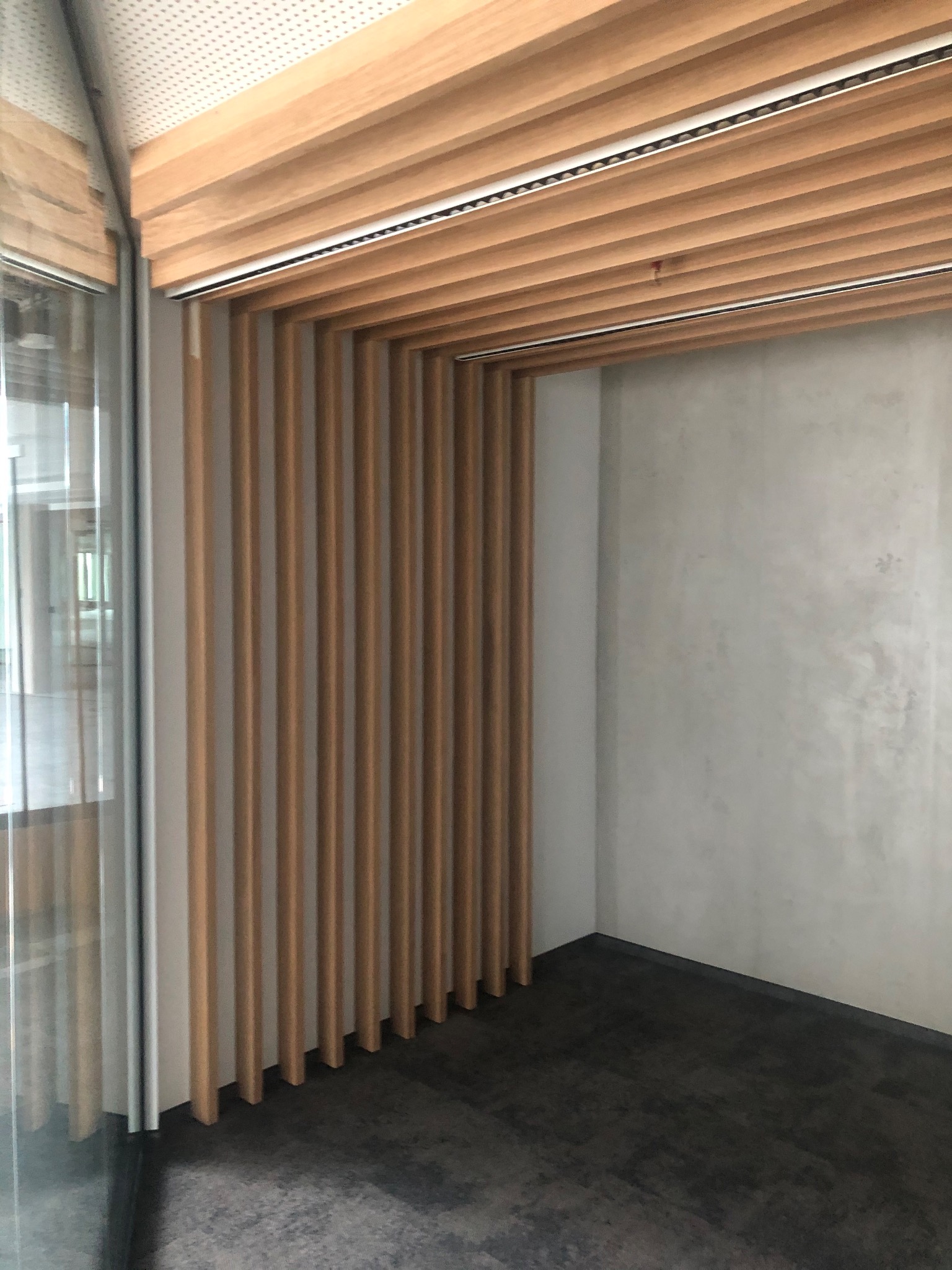 Nowoczesne wykończenie wnętrza biura z drewnianymi lamelami na suficie i ścianie, szara ściana z efektem betonu, ciemna podłoga i przeszklenie.