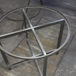 Metalowa konstrukcja stołu z okrągłym blatem w trakcie spawania w warsztacie, widoczne ślady obróbki metalu na podłodze.