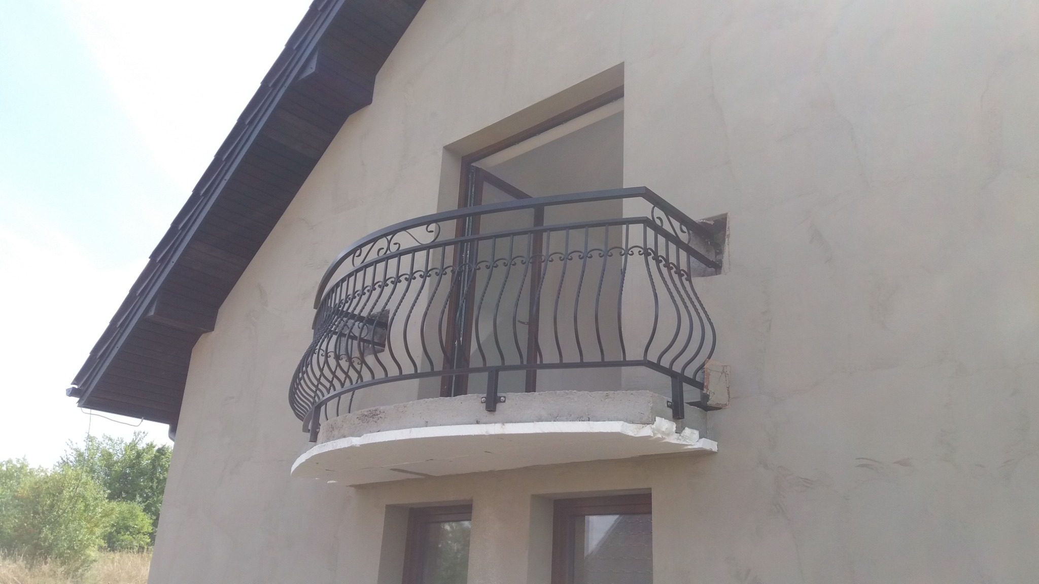 Kuty balkon z czarną balustradą zamontowany na budynku w trakcie wykańczania, widoczne niedoskonałości tynku i wystający styropian.