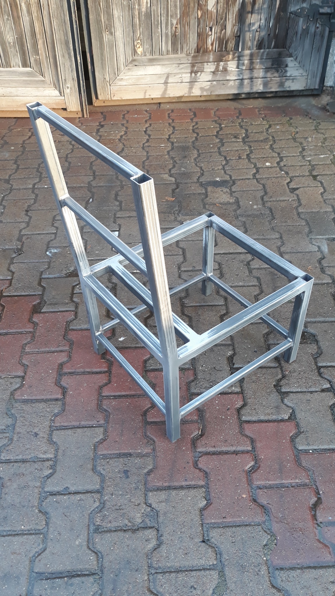 Metalowy szkielet krzesła wykonany z profili kwadratowych, spawany, widoczny na tle kostki brukowej i drewnianych drzwi.