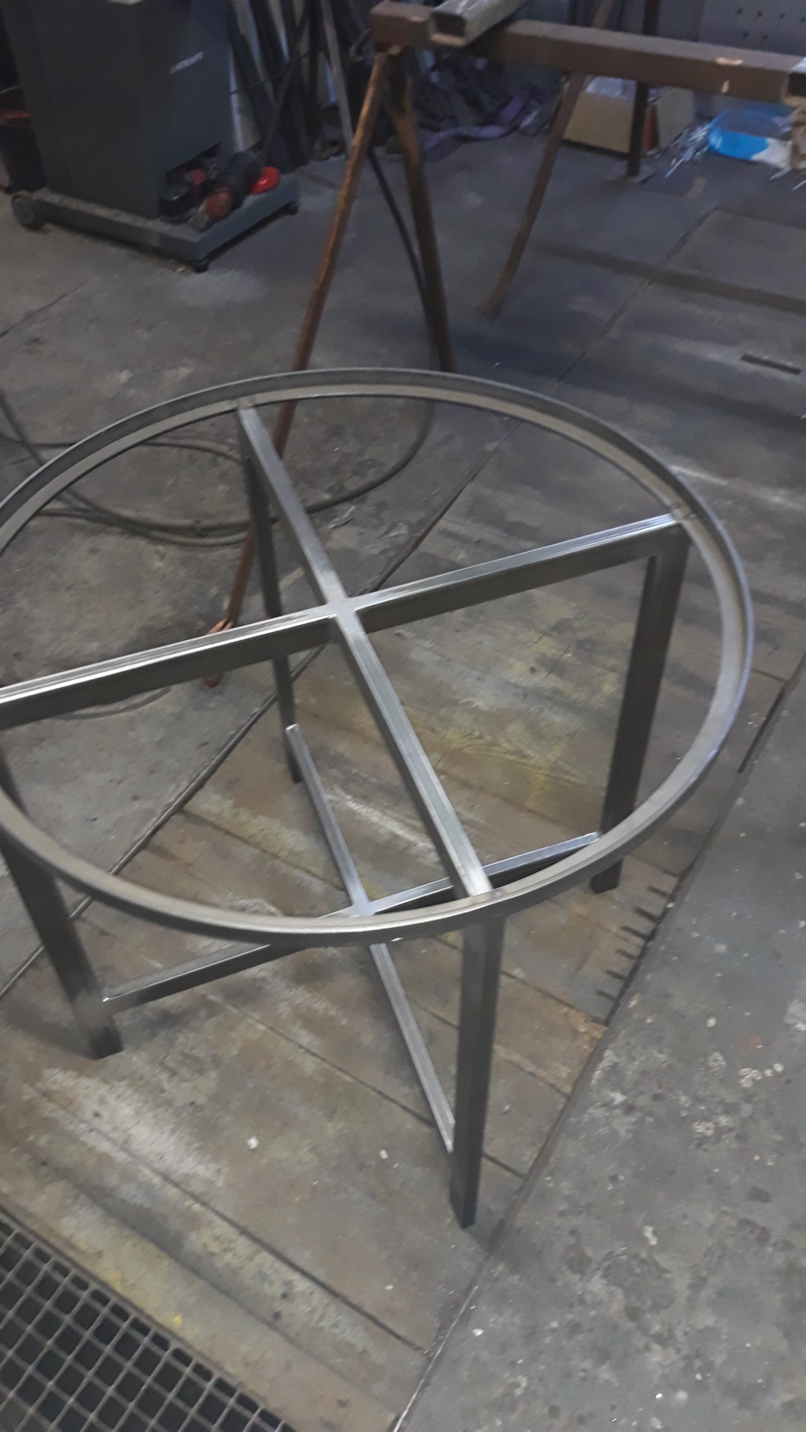 Metalowa konstrukcja stołu z okrągłym blatem w trakcie spawania w warsztacie, widoczne ślady obróbki metalu na podłodze.