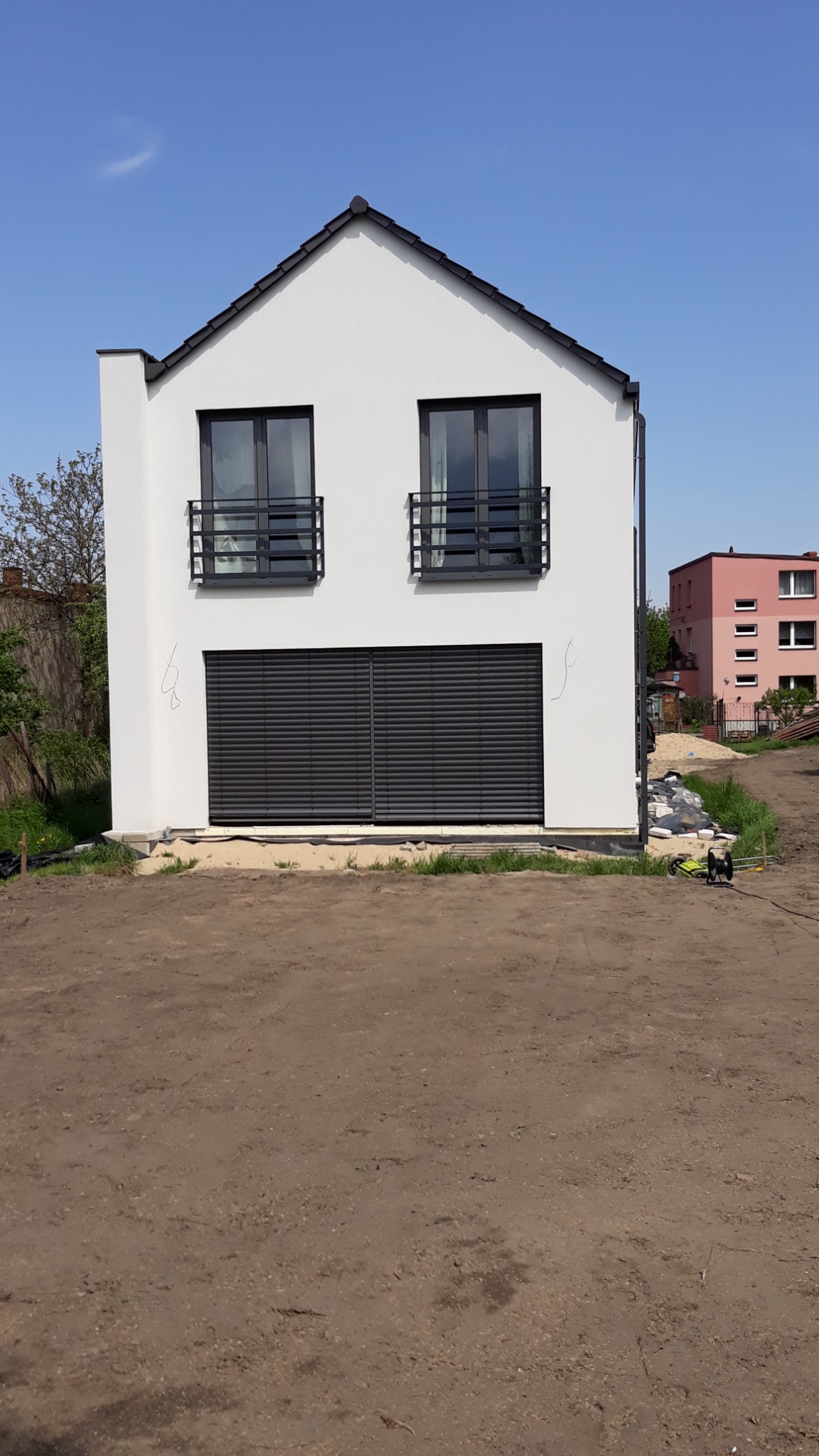 Nowoczesny, biały dom z czarnymi akcentami w Dąbrowie Górniczej, z widocznymi roletami zewnętrznymi i minimalistycznymi balustradami okiennymi, na tle niezagospodarowanej działki.