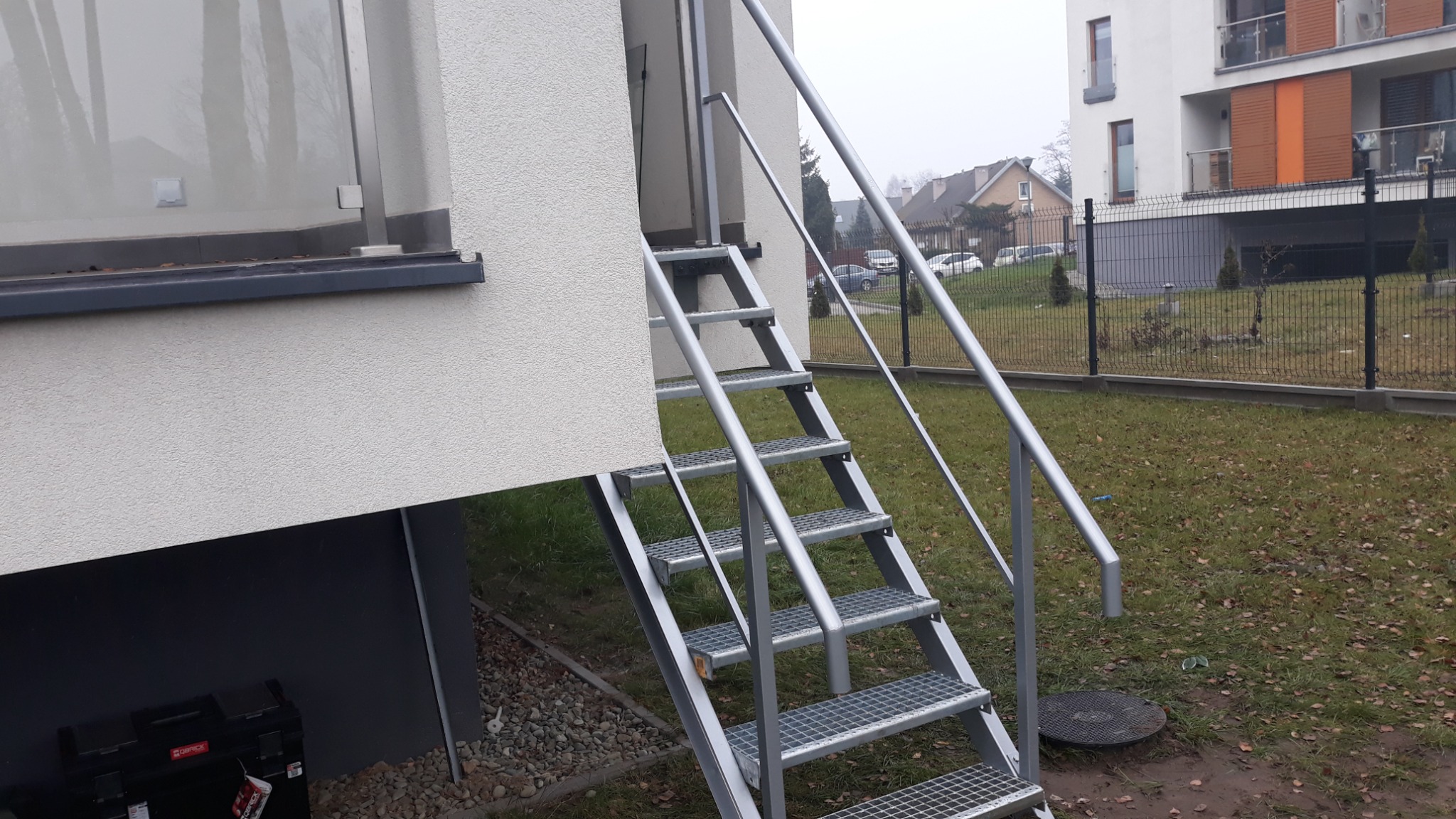 Zewnętrzne metalowe schody z kratowymi stopniami i poręczami prowadzące do wejścia w budynku, z widocznym fragmentem ogrodzenia panelowego w tle.