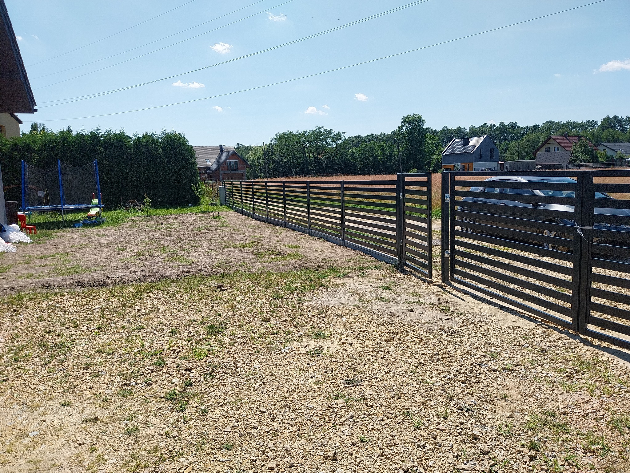 Nowoczesne, poziome ogrodzenie panelowe w kolorze antracytowym z bramą, oddzielające posesję z widocznym domem jednorodzinnym i trampoliną w tle.