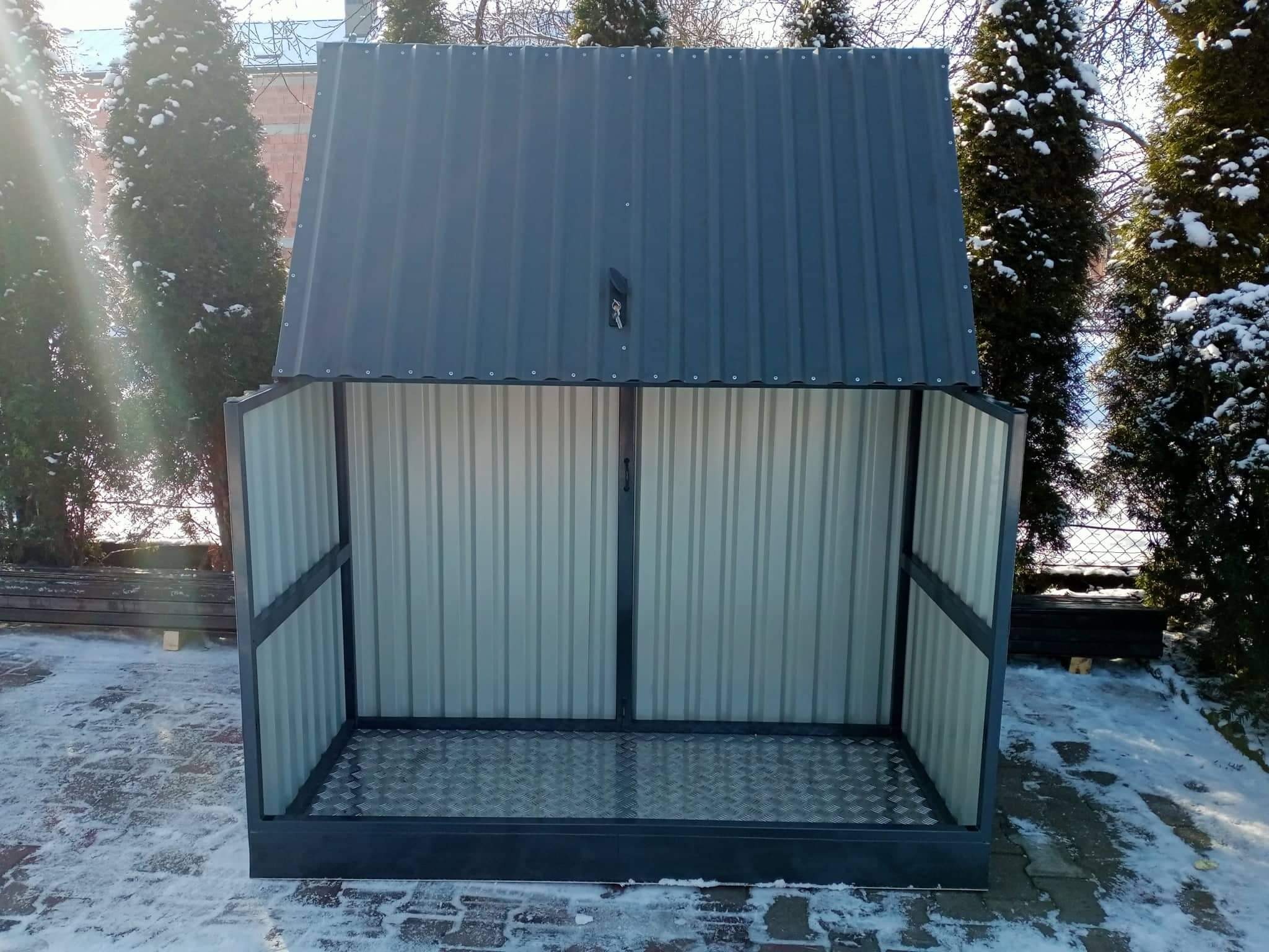 Metalowa konstrukcja z otwieraną górną częścią, przypominająca schowek lub mały garaż, z blachy falistej, umieszczona na zewnątrz na tle drzew pokrytych śniegiem.