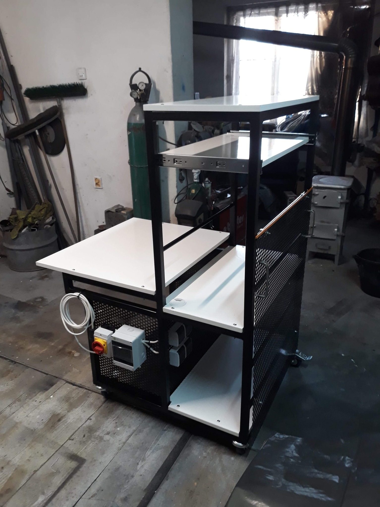 Wielopoziomowy wózek warsztatowy z czarną, spawaną konstrukcją i białymi półkami, wyposażony w kółka i perforowane ścianki boczne, widoczne przewody elektryczne i osprzęt, w tle butla z gazem...