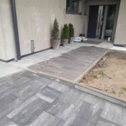 Nowa, szara kostka brukowa ułożona przed wejściem do domu, z obrzeżami i trzema tujami w donicach przy ścianie. Część terenu przygotowana pod dalsze prace.