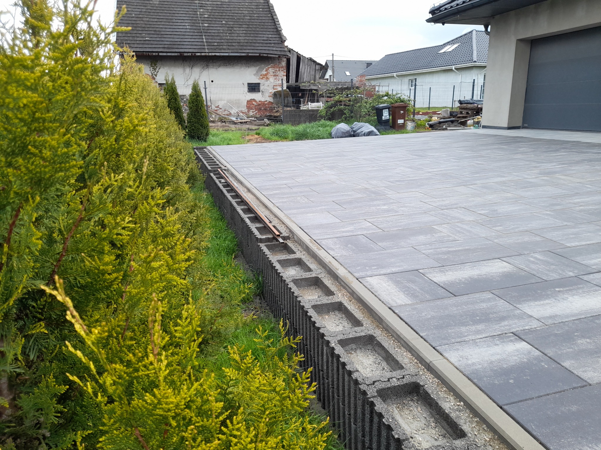 Szara kostka brukowa ułożona na podjeździe, obrzeża z betonowych bloczków, w tle budynek gospodarczy w stanie rozpadu i nowoczesny dom z garażem.