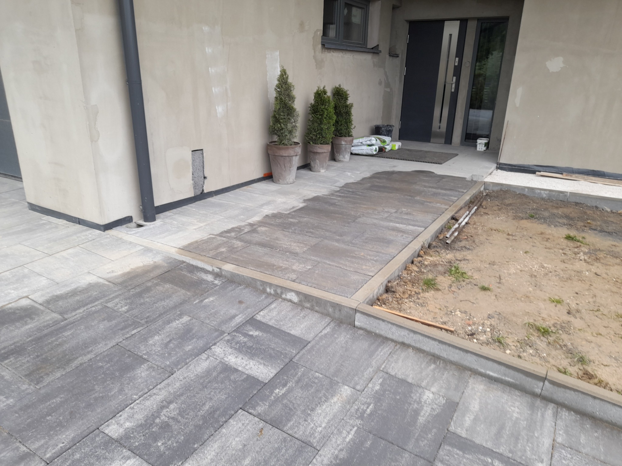 Nowa, szara kostka brukowa ułożona przed wejściem do domu, z obrzeżami i trzema tujami w donicach przy ścianie. Część terenu przygotowana pod dalsze prace.