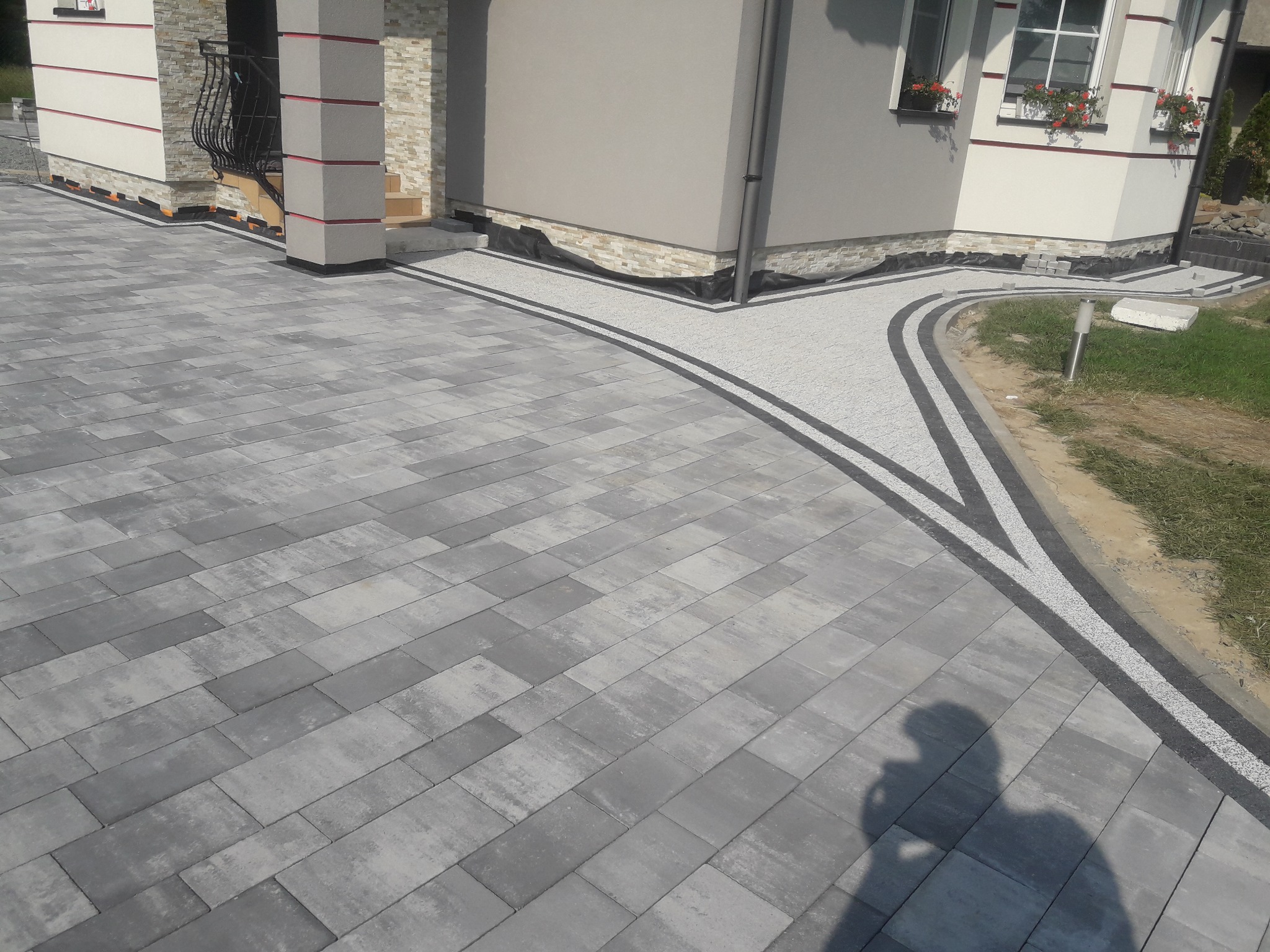 Ułożona szara kostka brukowa przed domem z ozdobnymi pasami z jasnego kruszywa, widoczny cień osoby fotografującej.