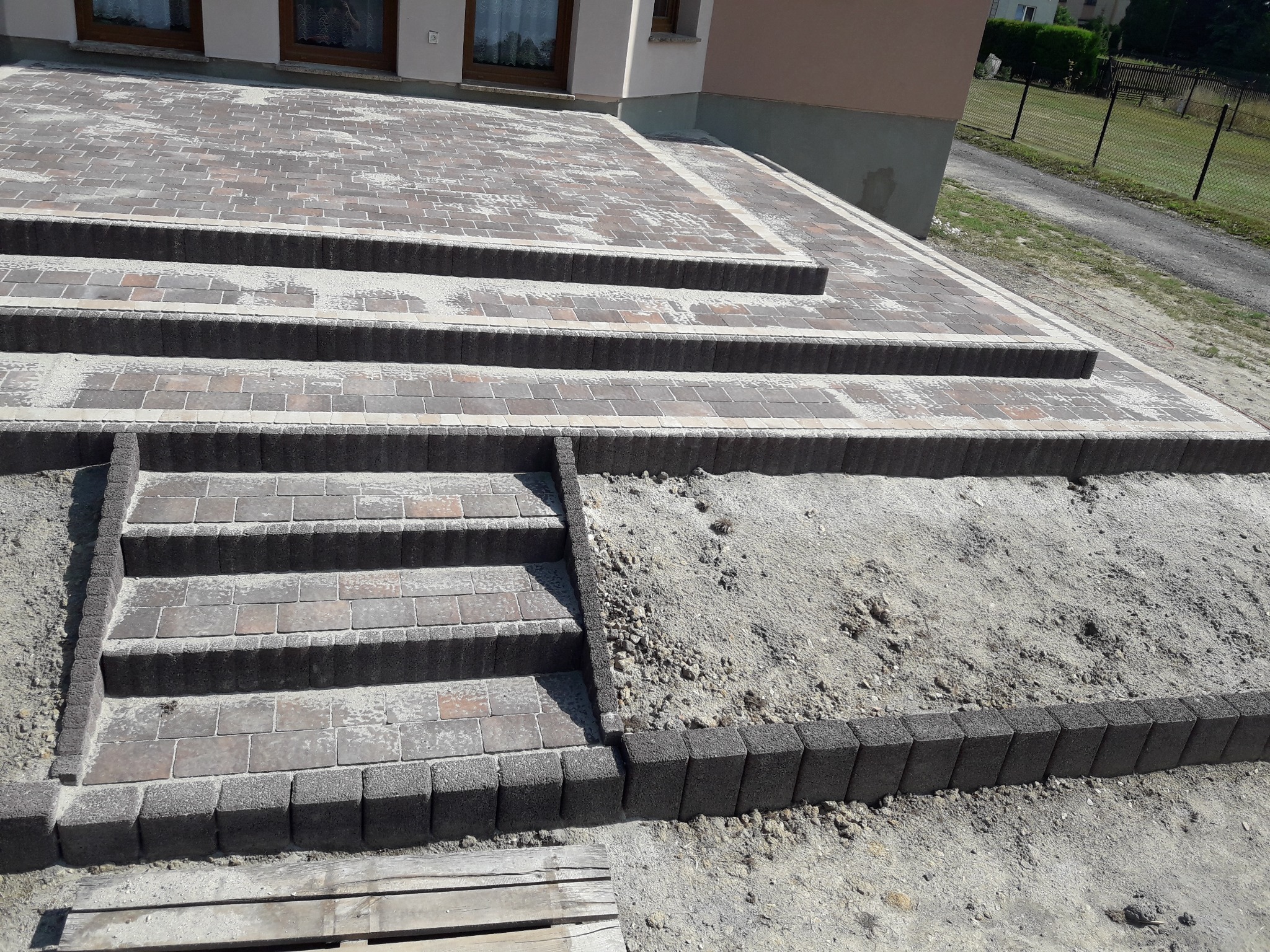 Schody i podjazd z kostki brukowej w odcieniach szarości i brązu, częściowo zasypane piaskiem, widoczne elementy drewniane w dolnym rogu.